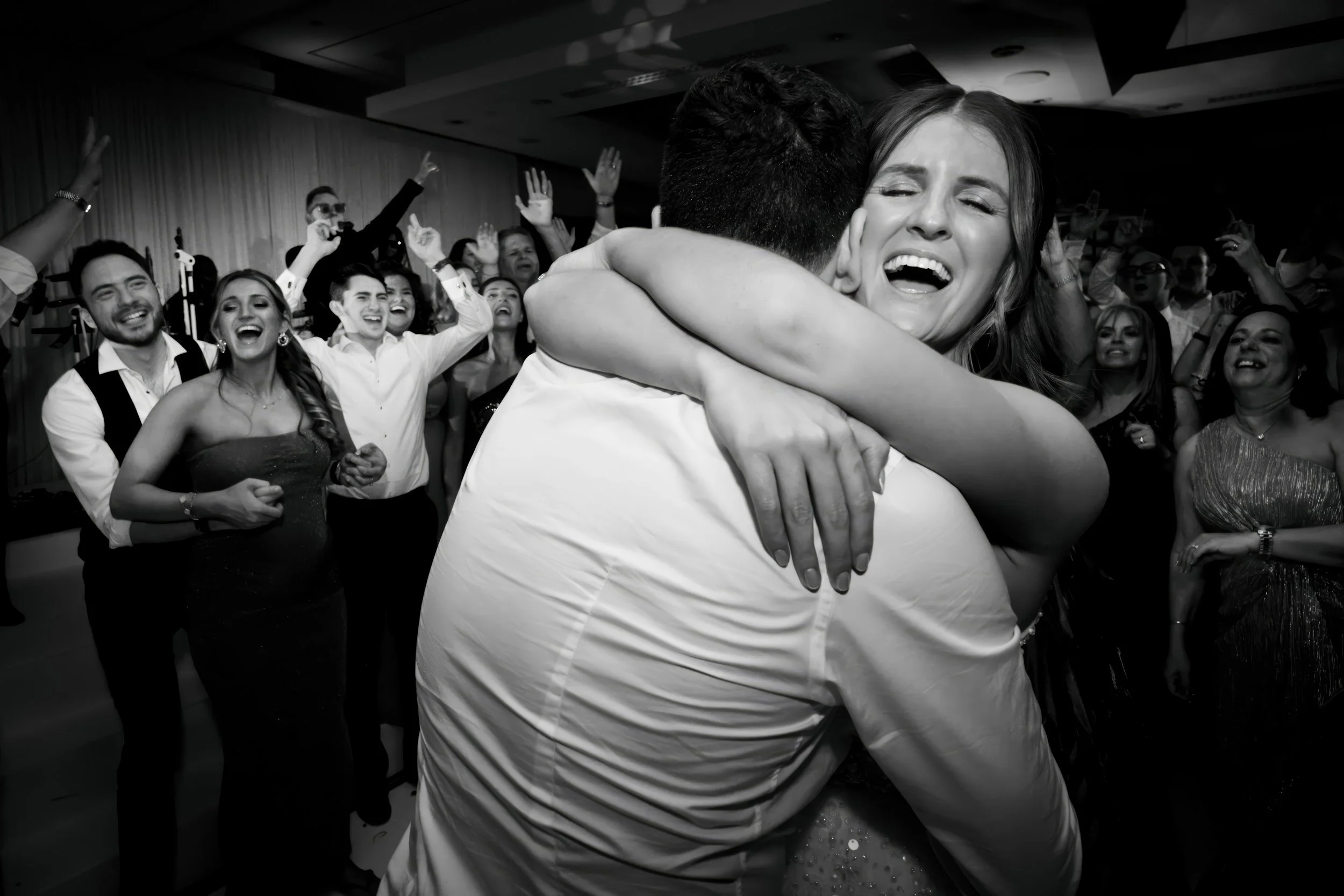 A woman and a man hugging in a dance floor while a group of people look on, celebrating at a party or wedding reception.
