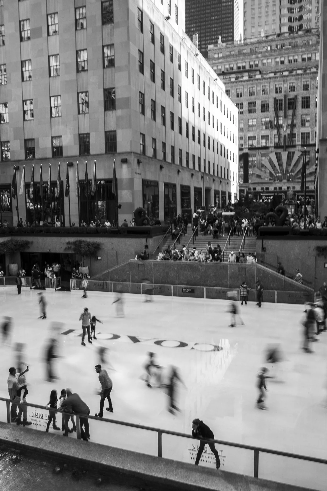 Black and white photo of the Rockefeller ice skating rink in New York City with people skating and sitting around.