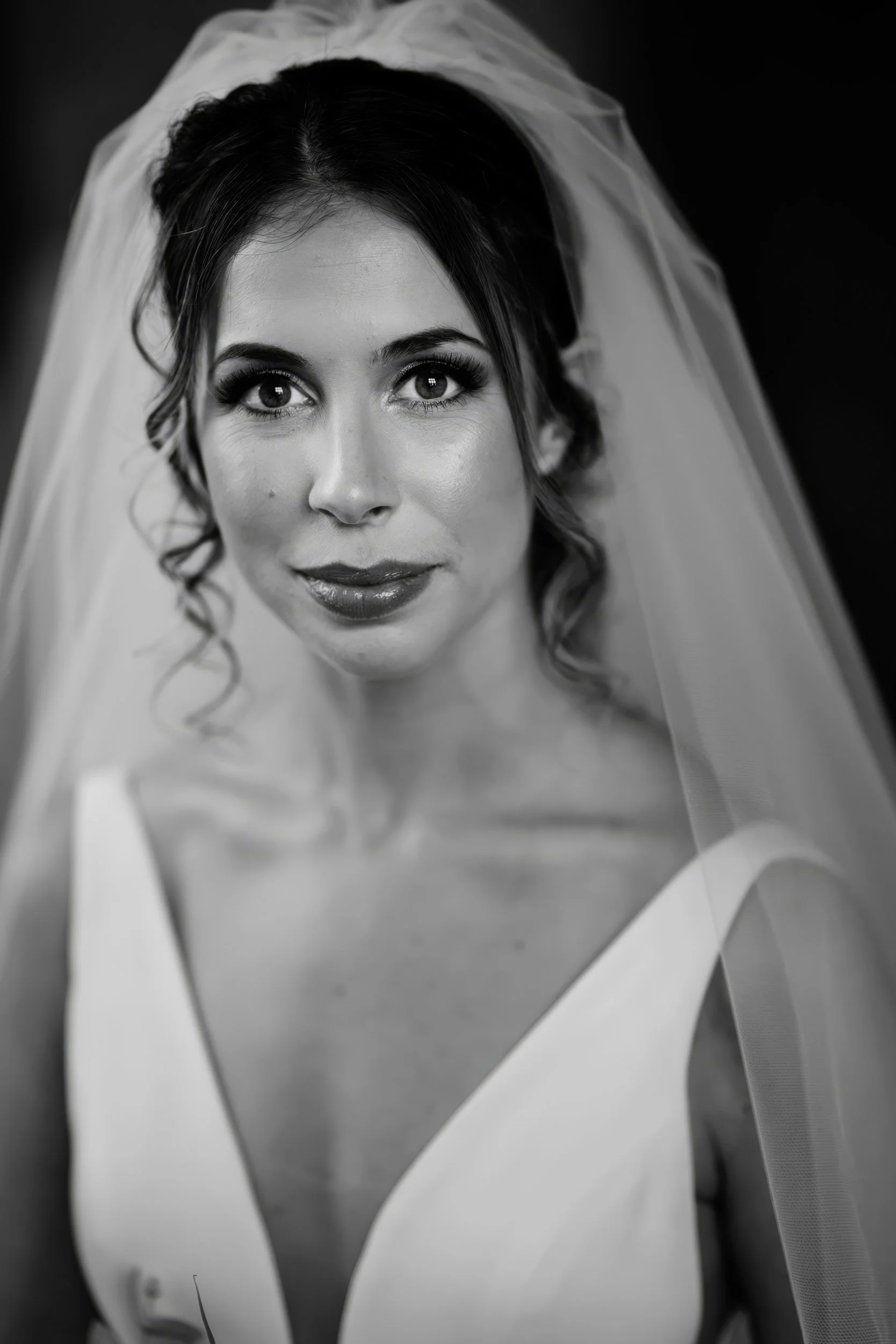 Close-up black and white portrait of a bride with a veil, wearing a sleeveless dress, looking directly at the camera with a gentle smile.