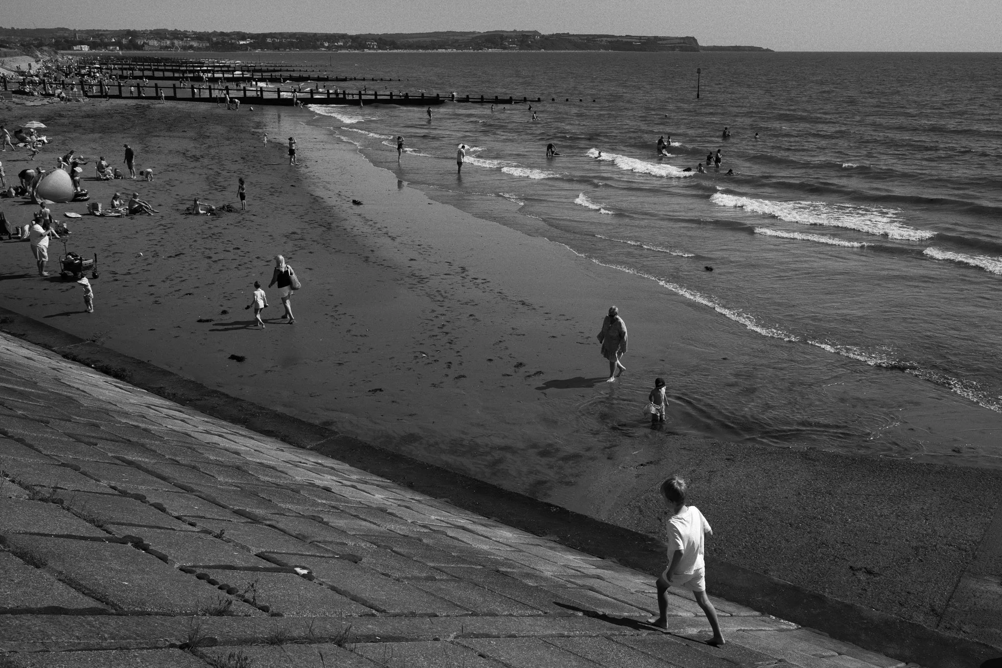 Beach scene with people walking along the shore, swimming, and relaxing, taken from an elevated vantage point, with a pier extending into the water in the background.