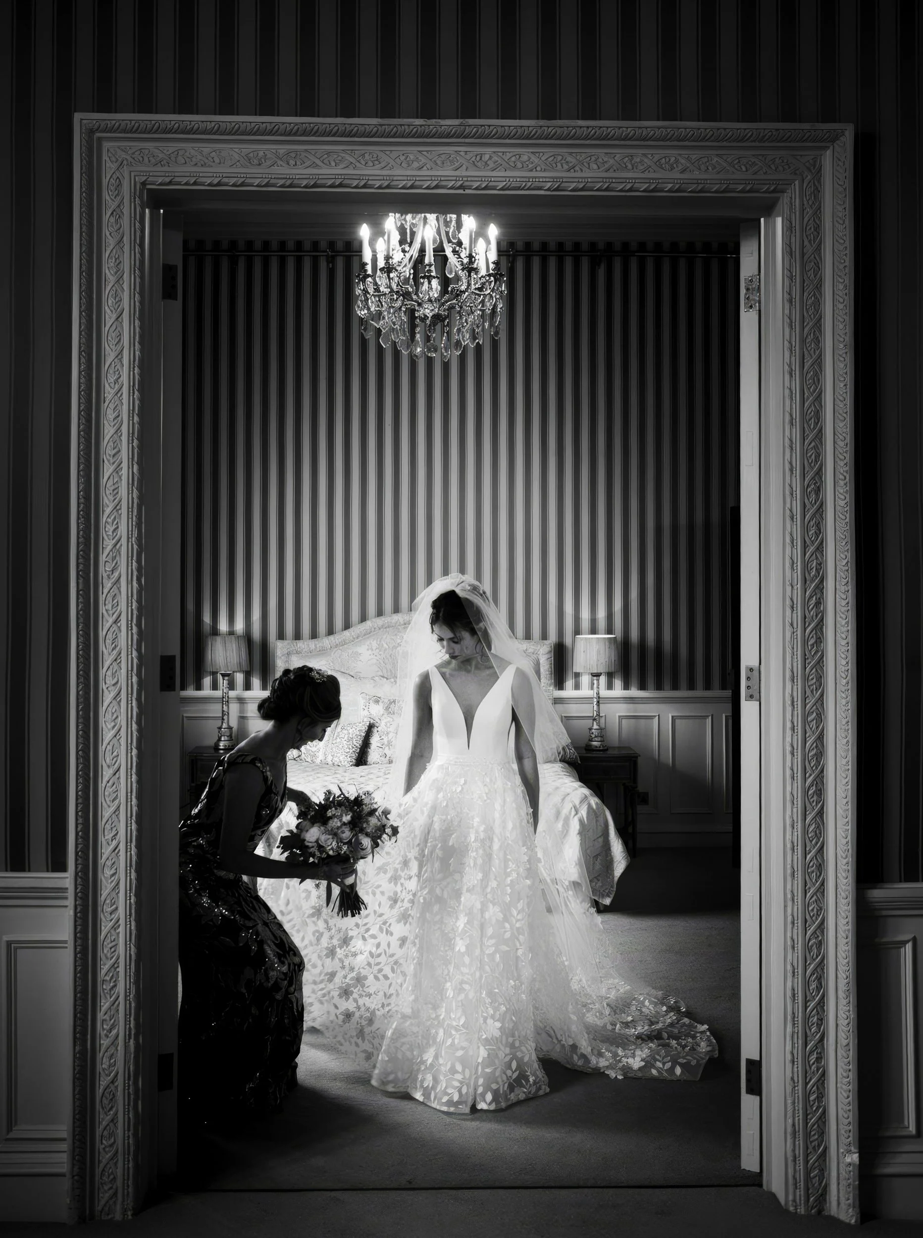 A bride in a wedding gown with veil inside a bedroom, with a woman kneeling, holding a bouquet of flowers, helping her prepare.