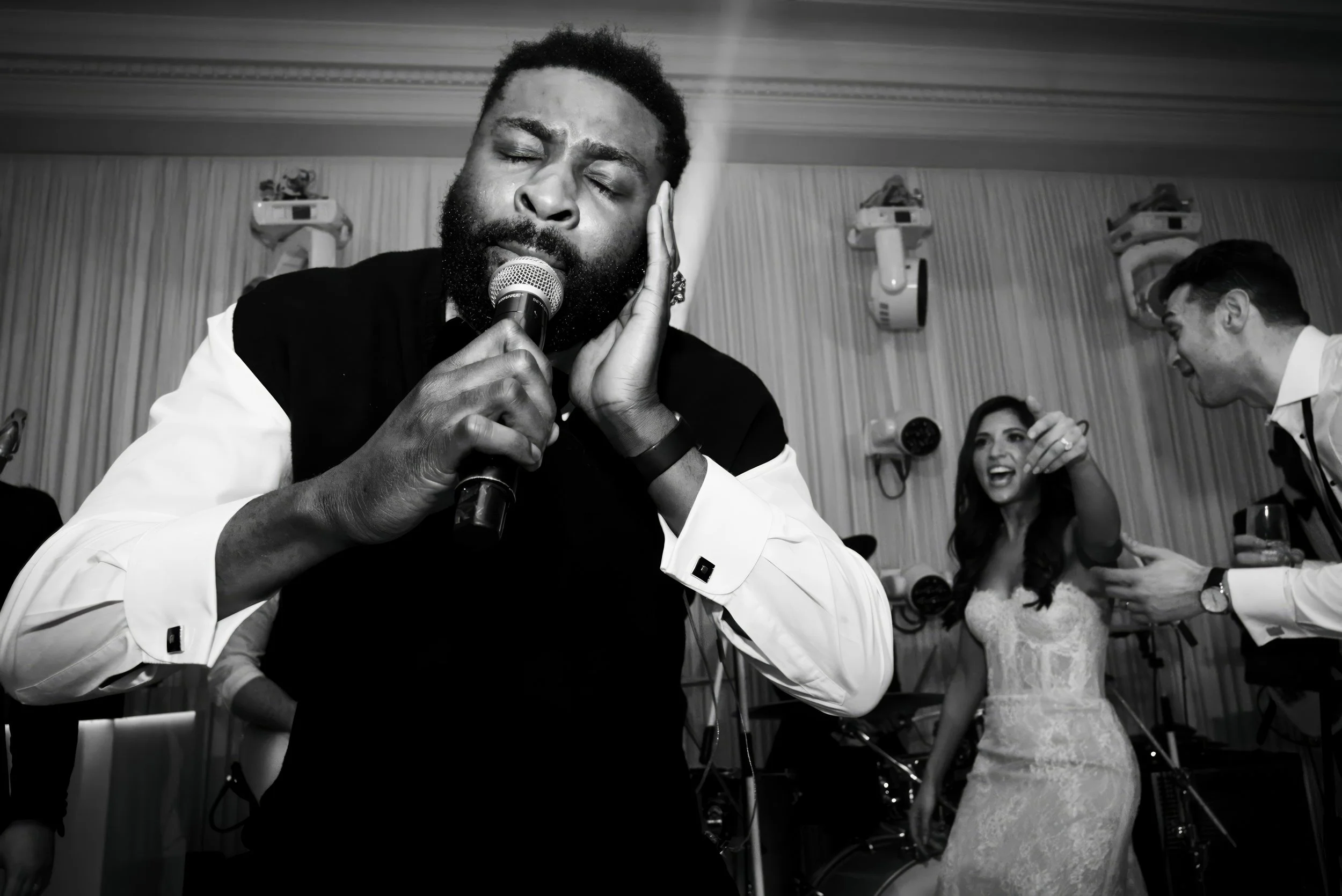 A man singing passionately at an event, holding a microphone with eyes closed and hand on his face, while a woman in a wedding dress and a man in a tuxedo dance and smile in the background.