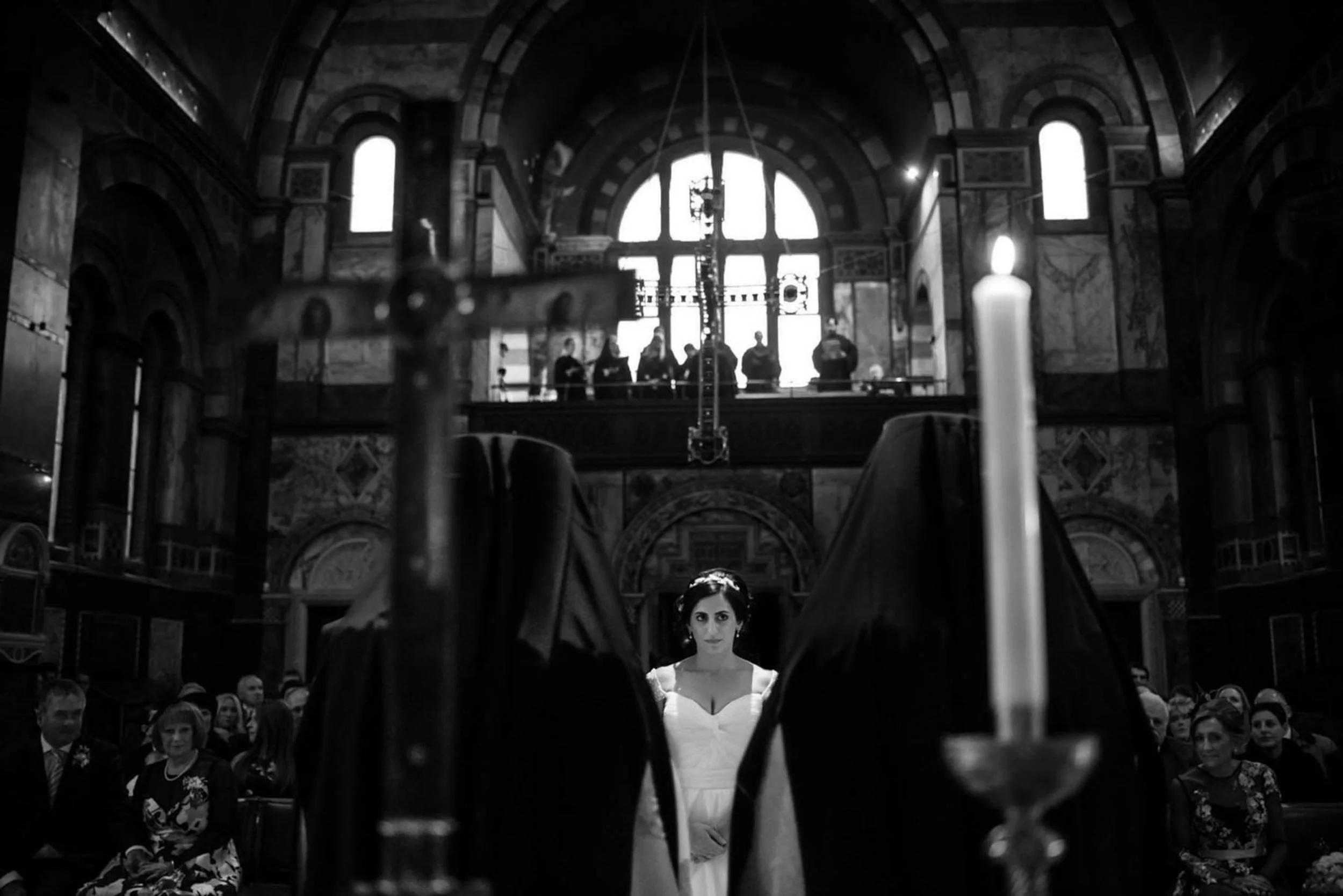 Black and white photo of a woman in a wedding dress standing in a cathedral with guests seated on either side. Candles and religious artifacts are in the foreground, with a group of people on a balcony in the background.