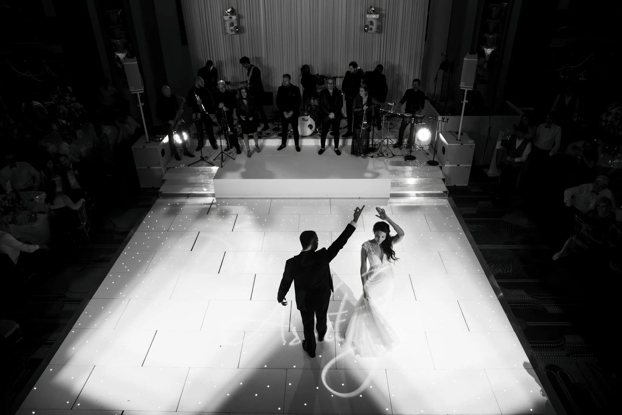 Black and white photo of a bride and groom dancing on a lit dance floor, with a live band and guests watching in the background at a wedding reception.