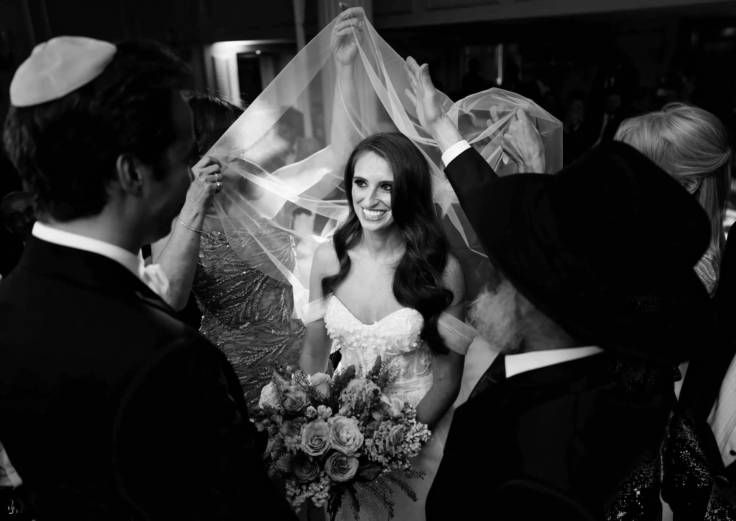 Black and white photo of a bride with long dark hair smiling under a veil held by surrounding people at her wedding.