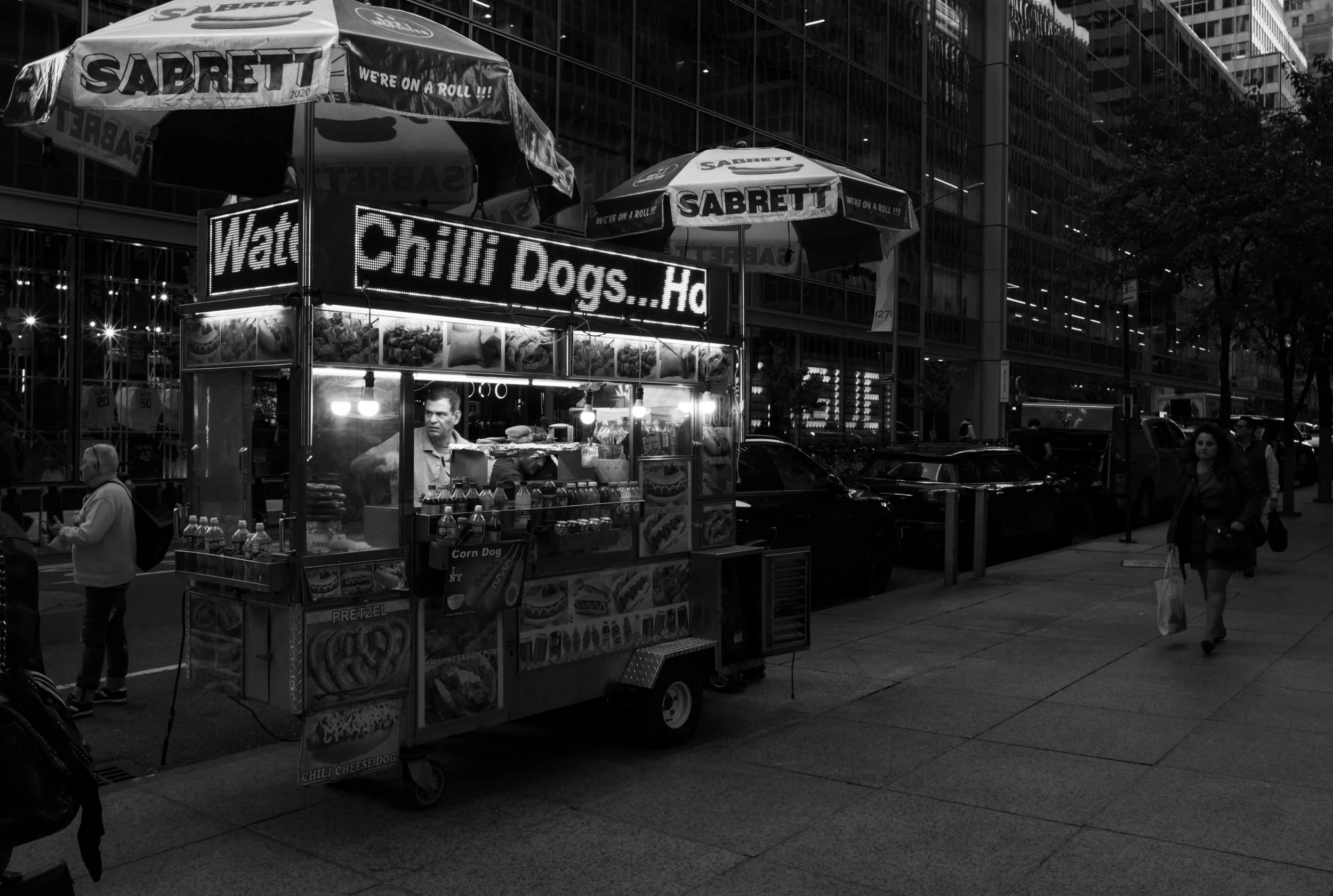 A hot dog stand on the sidewalk in an urban area at night with a brightly lit sign advertising hot dogs and other snacks, surrounded by parked cars and walking pedestrians.