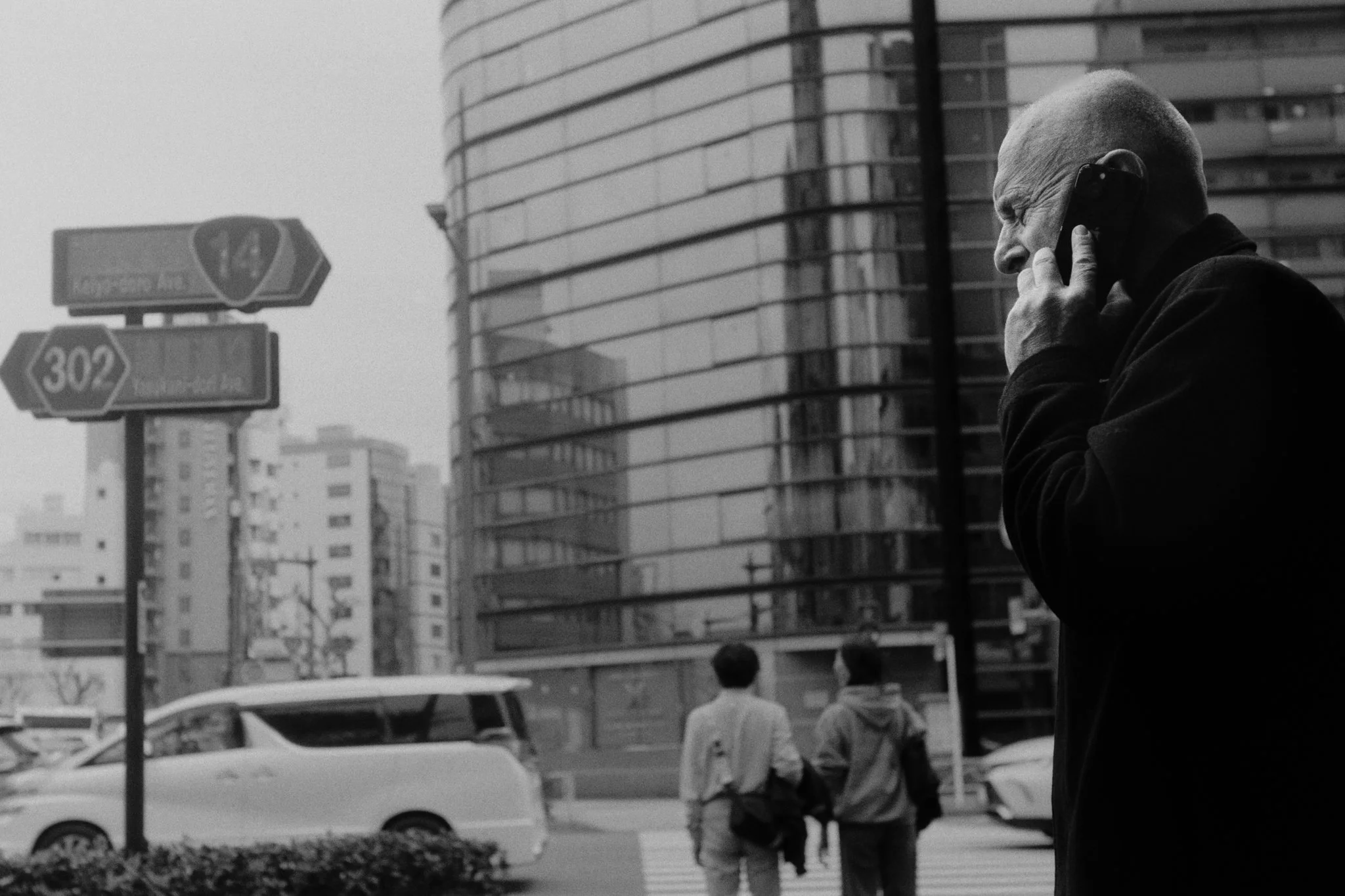 A man with a shaved head shown in profile, talking on a cellphone on a city street with modern buildings and cars, two people crossing the street, black-and-white photograph.