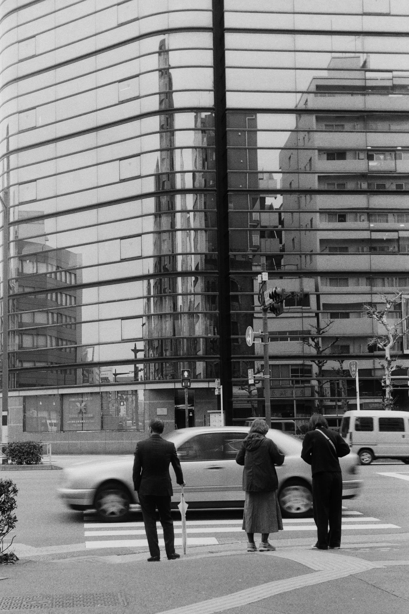 Three people waiting to cross the street in front of a modern glass building with reflections of other buildings, with cars passing by.