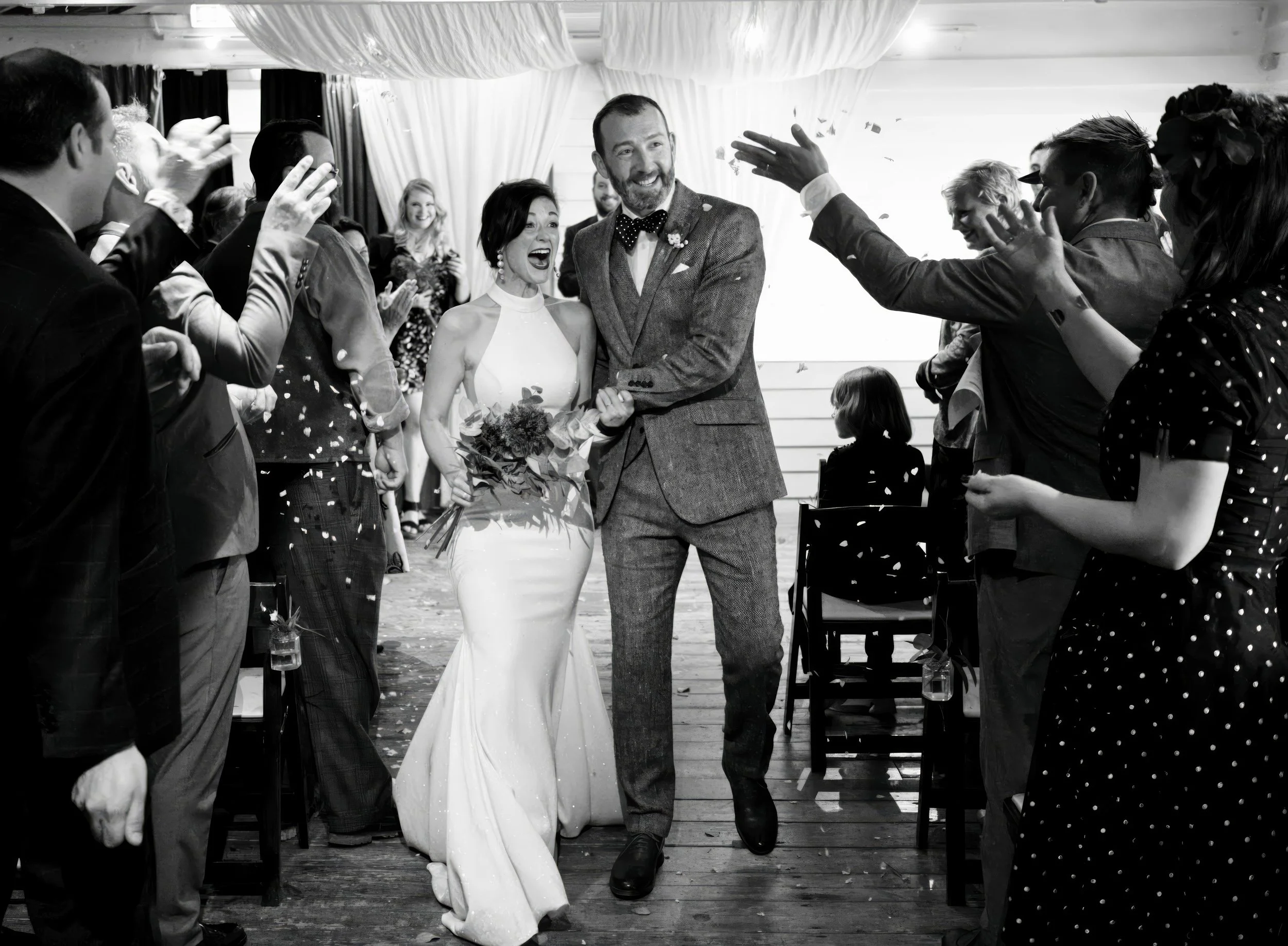 Black and white photo of a wedding celebration showing a bride and groom walking down the aisle. The bride is holding a bouquet and smiling, while the groom is holding her hand and smiling. Guests on both sides are clapping and cheering.