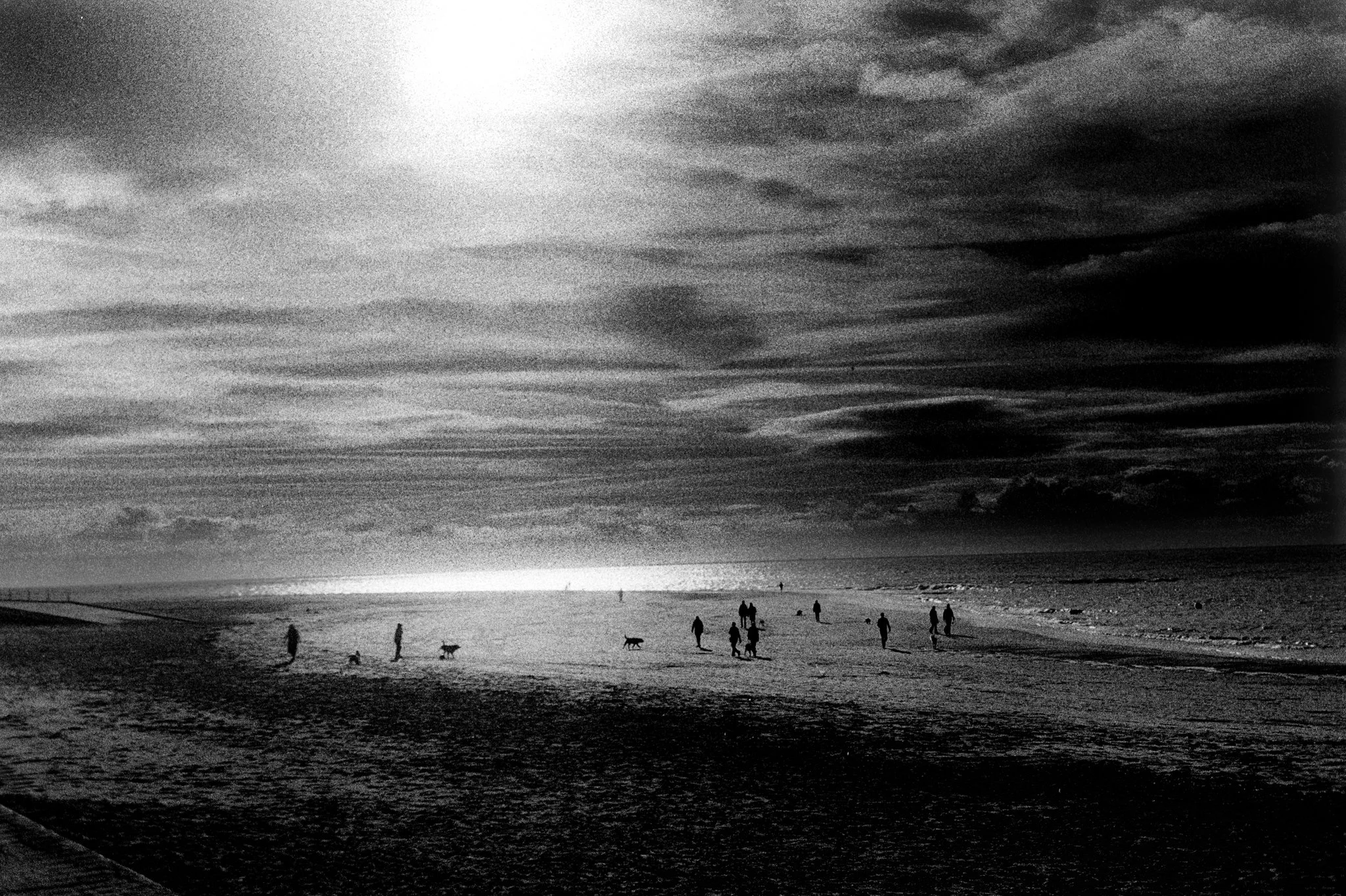 Black and white photo of a beach with several people walking and playing, and clouds in the sky.