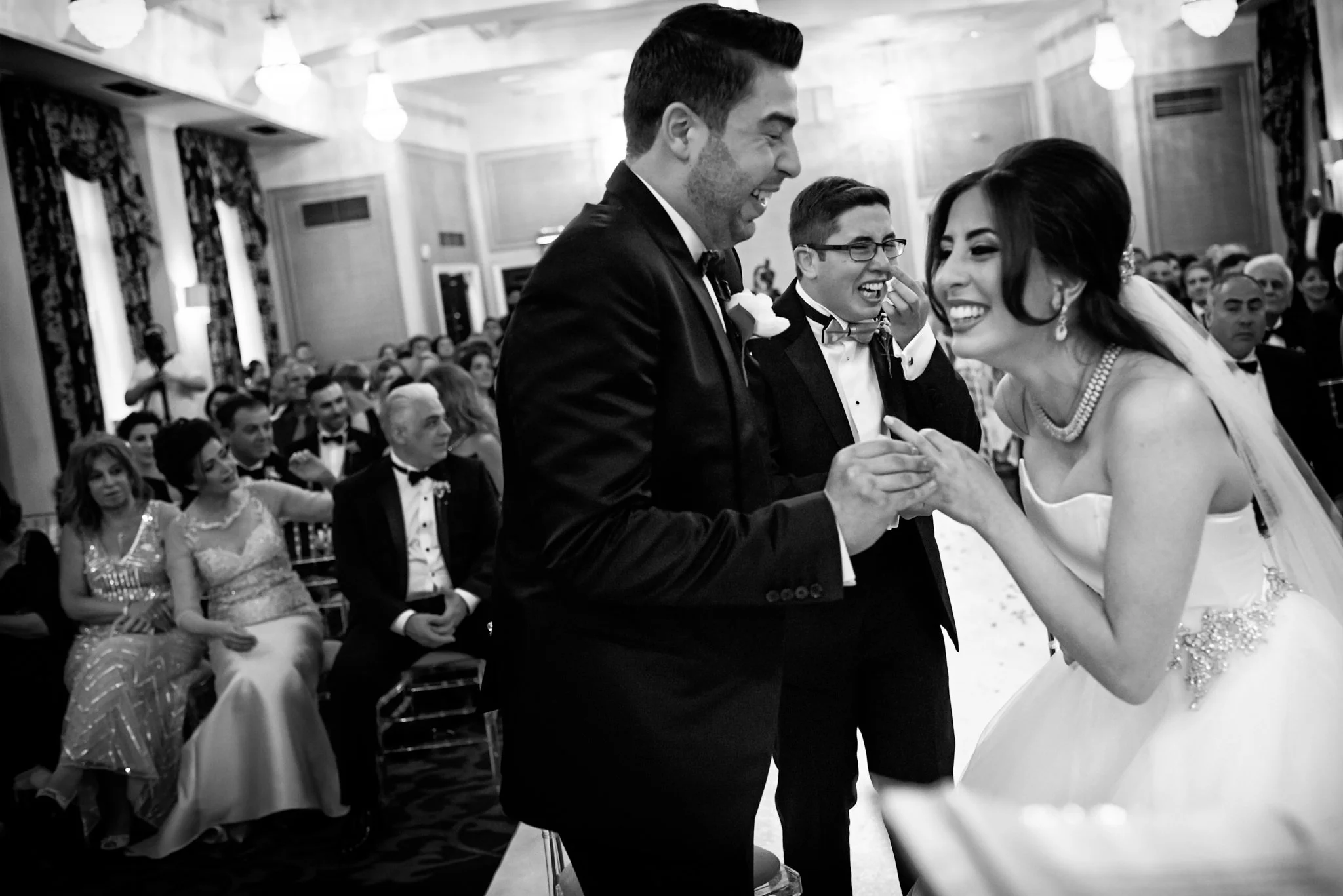A joyful wedding ceremony with a bride and groom exchanging rings, surrounded by guests in formal attire, in an elegant decorated hall.