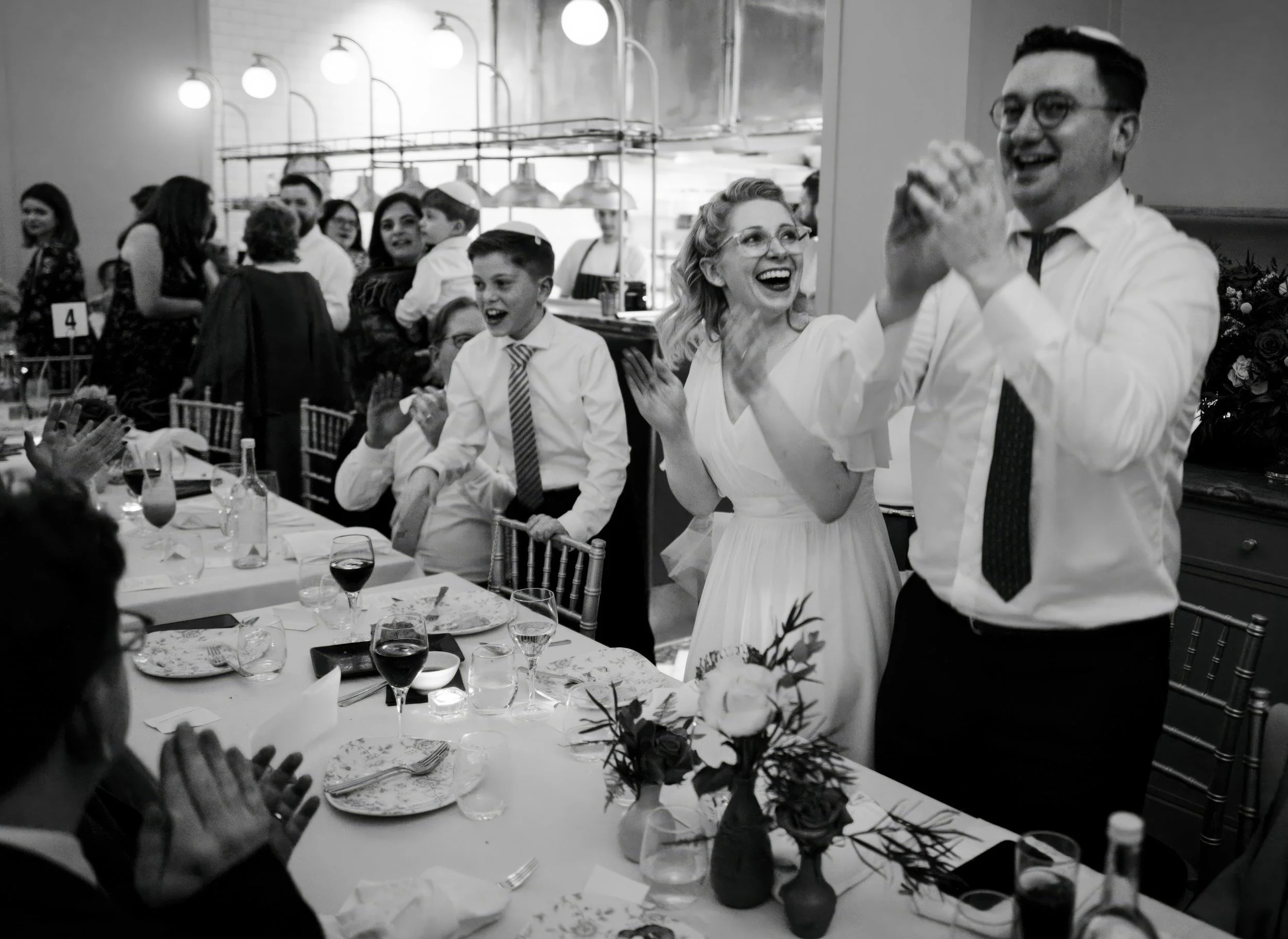 People celebrating at a wedding reception, including a woman and man dancing, with other guests clapping and talking in the background. The table has wine glasses, plates, and floral centerpieces.
