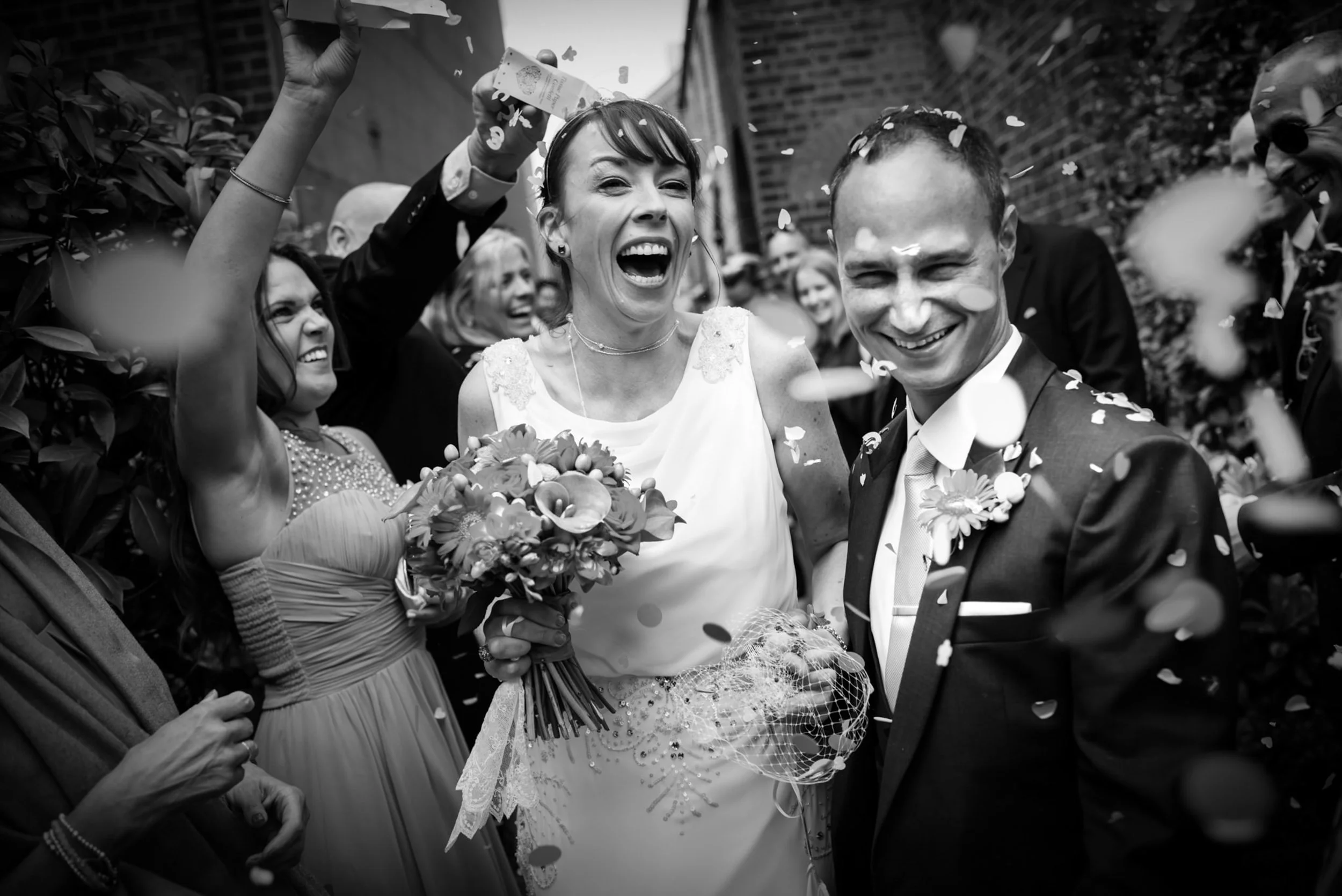 A black-and-white photo of a wedding celebration showing a happy bride and groom surrounded by friends and family. The bride holds a bouquet of flowers, laughing joyfully, while the groom smiles. Confetti is falling as they celebrate.
