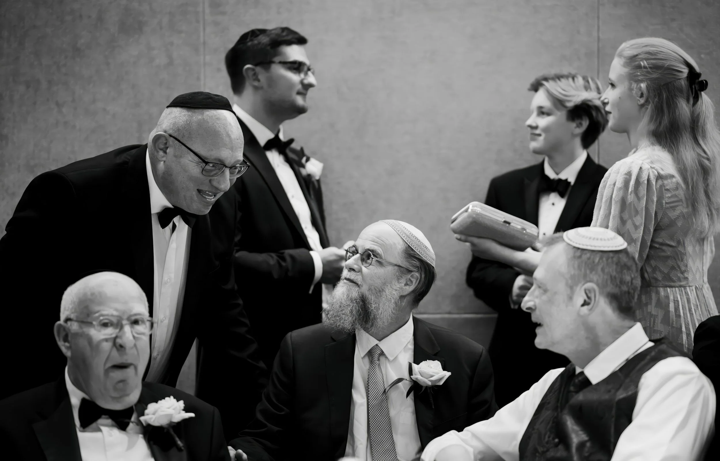 Group of Jewish men and women at a celebration, dressed in formal attire with yarmulkes, some with flowers on their suits, interacting and smiling, in a social setting.