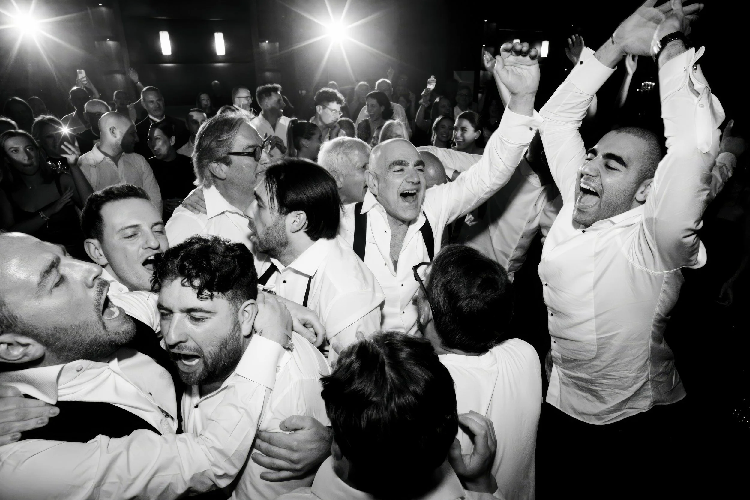 People celebrating and dancing at a party, smiling and enjoying themselves, with some individuals with their hands raised, in a lively and joyful atmosphere.