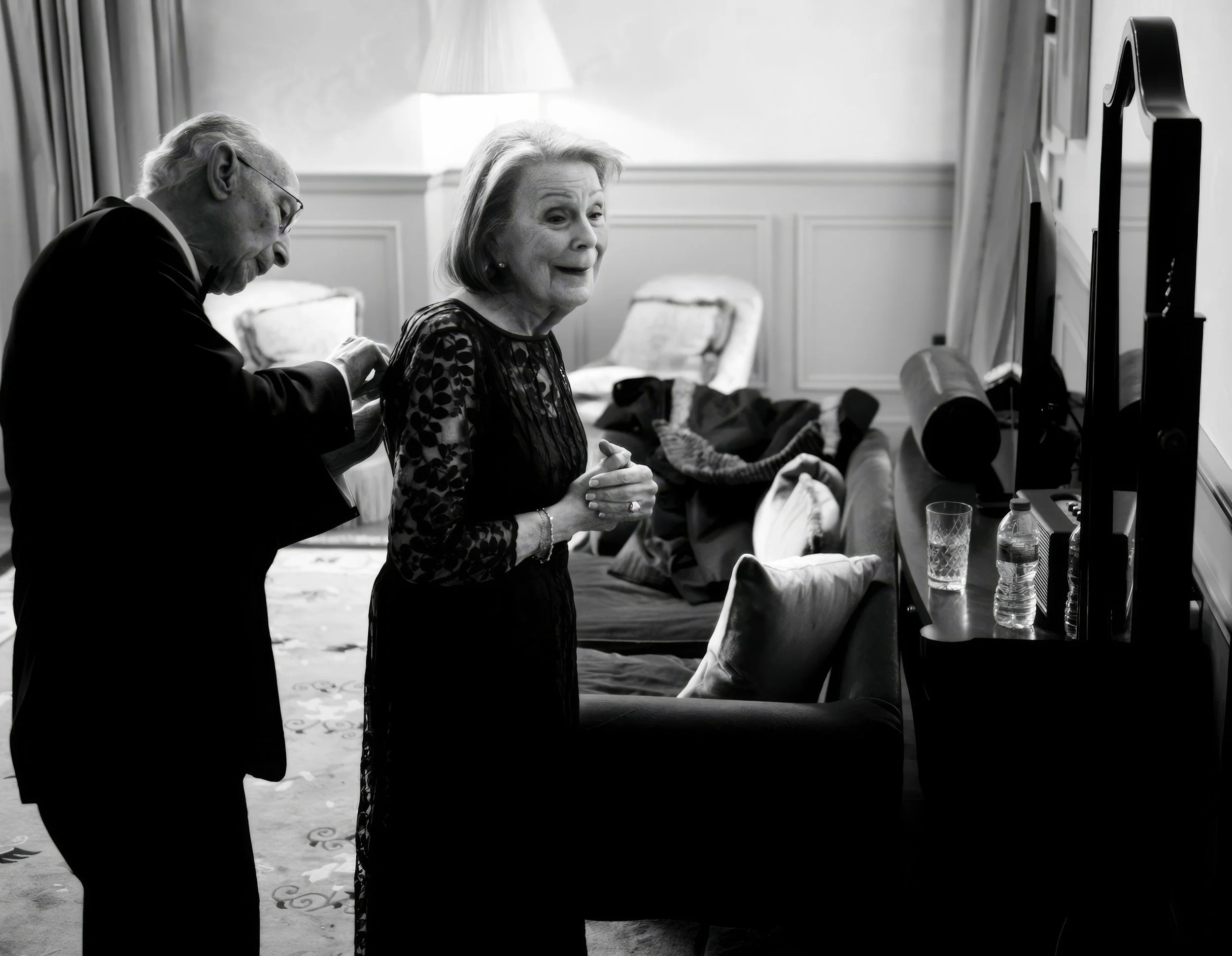 An elderly man and woman in a living room, with the man adjusting his jacket and the woman looking at the camera. The room has a TV, water bottles, glasses, and a sofa with blankets and pillows.