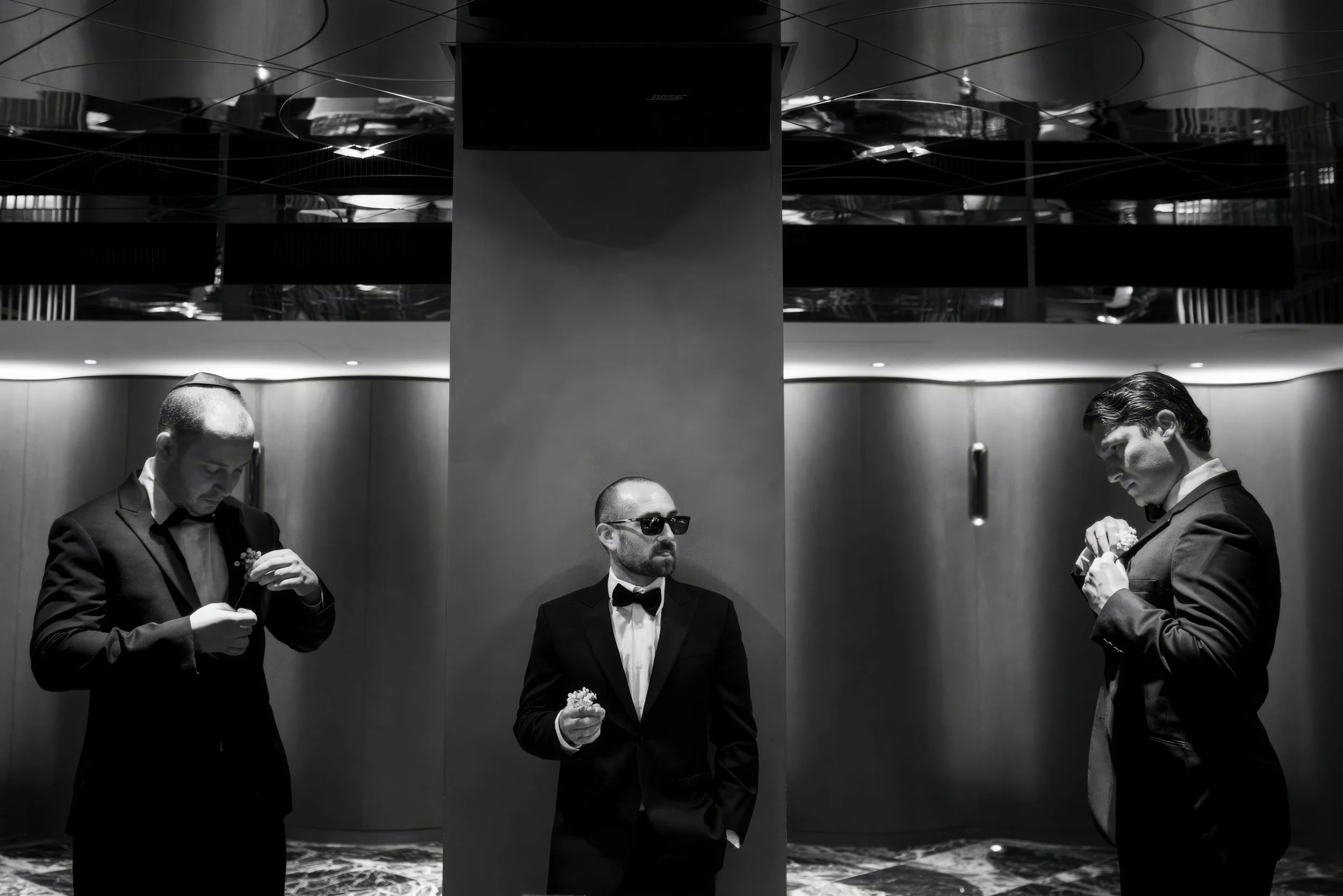 Three men in formal suits inside an elevator, with two of them examining objects and one man wearing sunglasses, all in black and white.