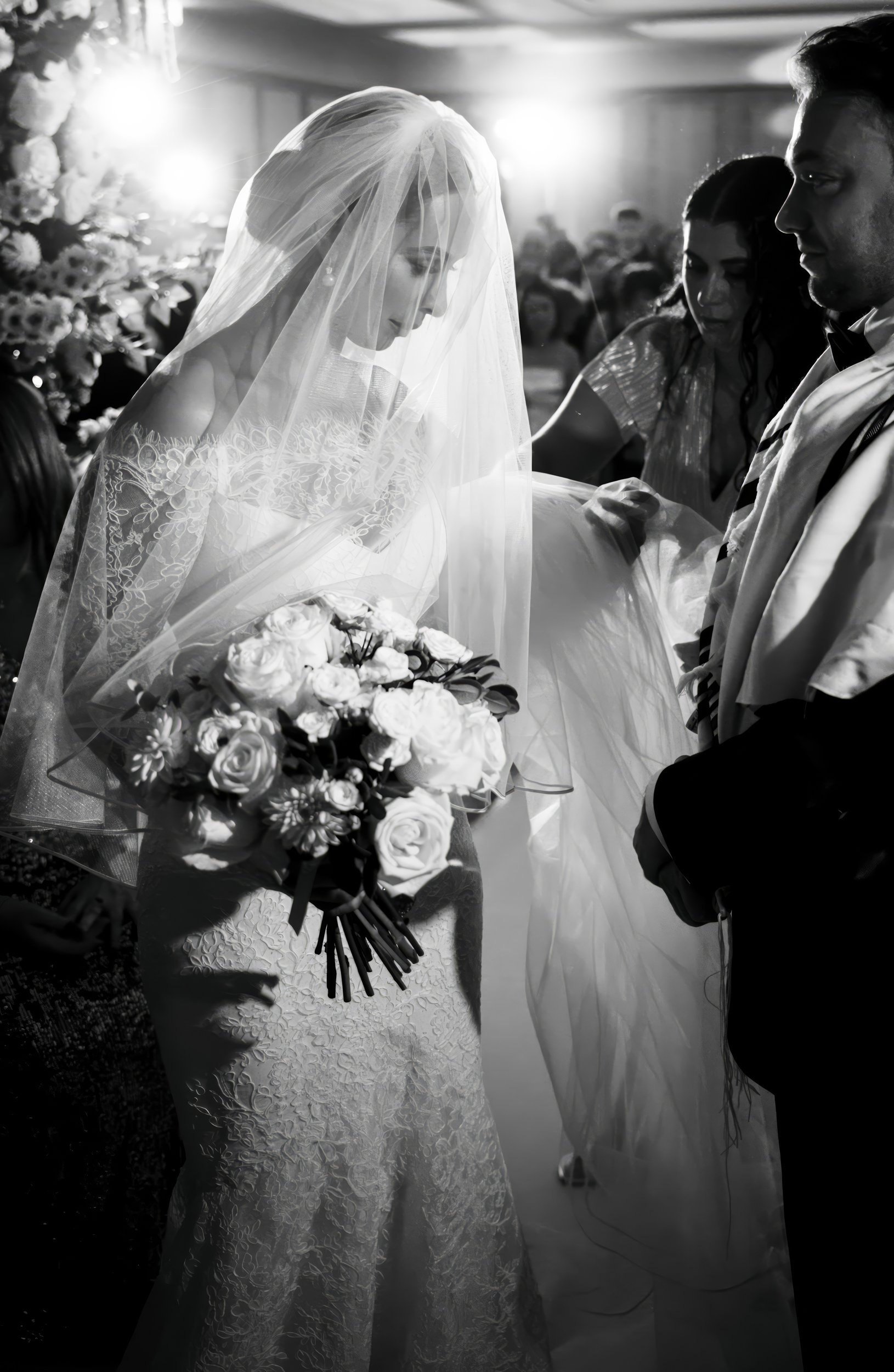 Black and white photo of a wedding scene showing a bride holding a bouquet of roses and other flowers, a gown, a veil, and people around her.