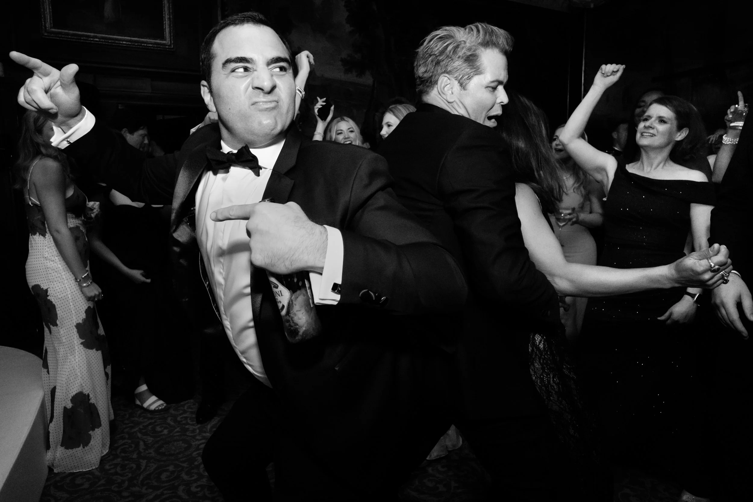 People dancing and enjoying themselves at a formal event, with men in tuxedos and women in elegant dresses, in a lively party atmosphere.