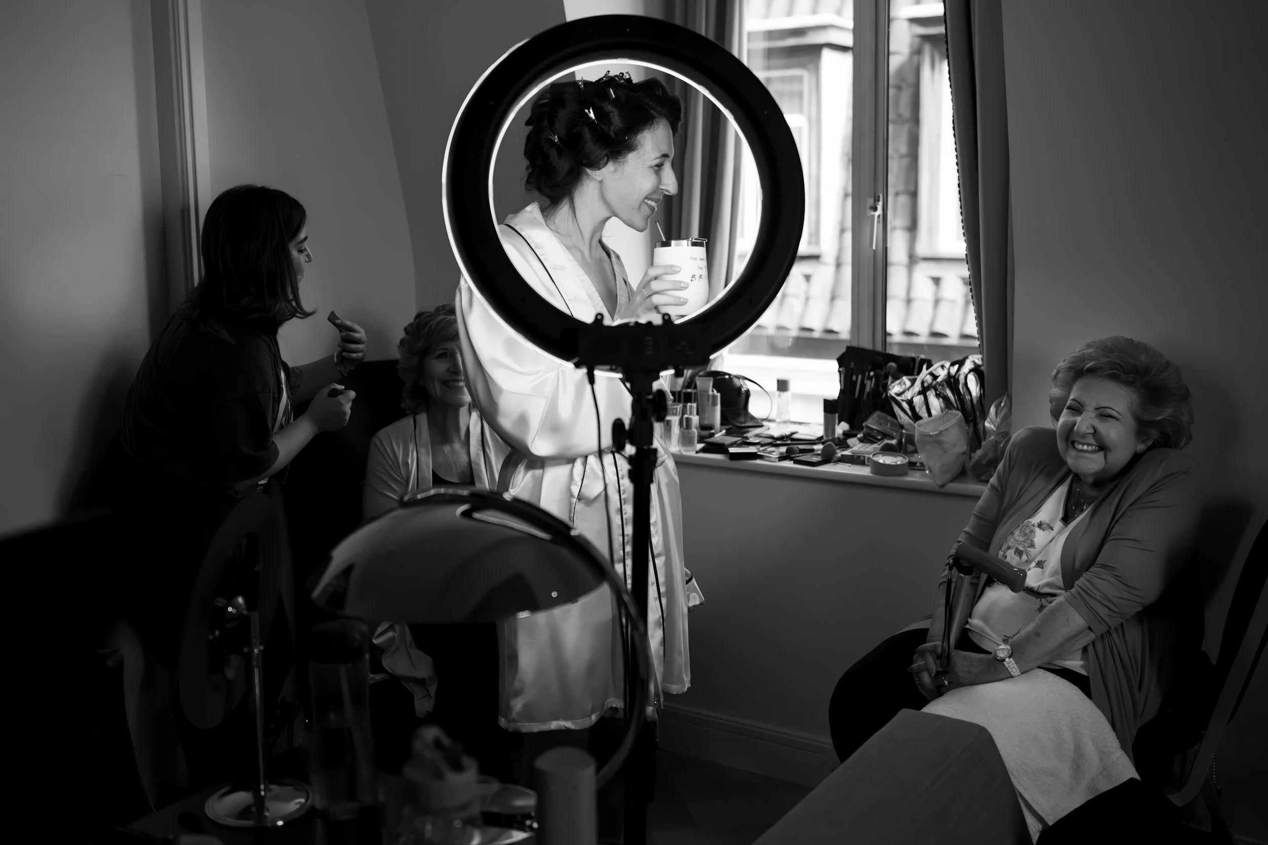 Black and white photo of a woman getting her makeup done while another woman attends to her, and a third woman sitting and smiling, in a room with makeup supplies on the windowsill. A large ring light on a stand is in the foreground.