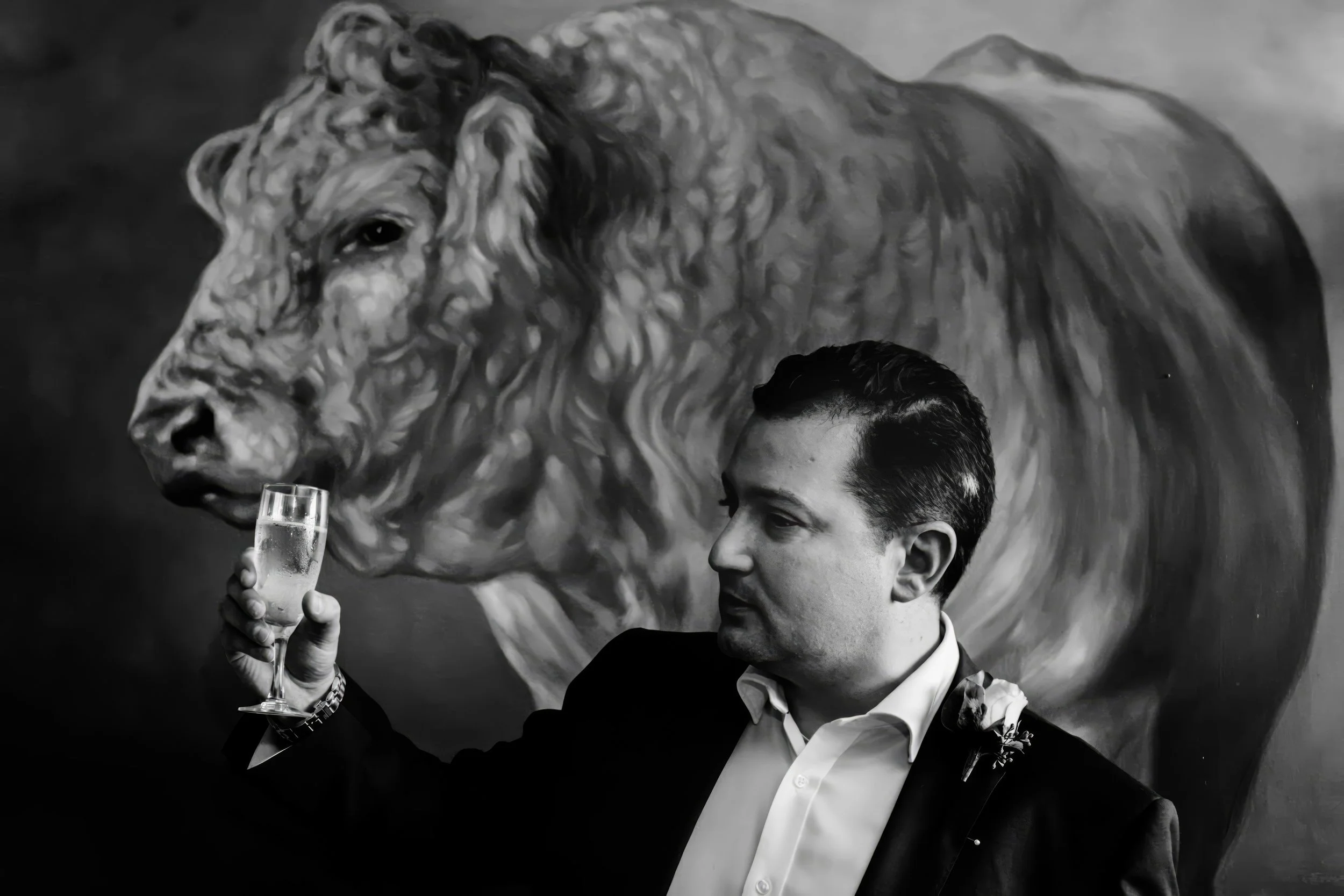 A man in a suit holding a glass of champagne, standing in front of a large painting of a lion.