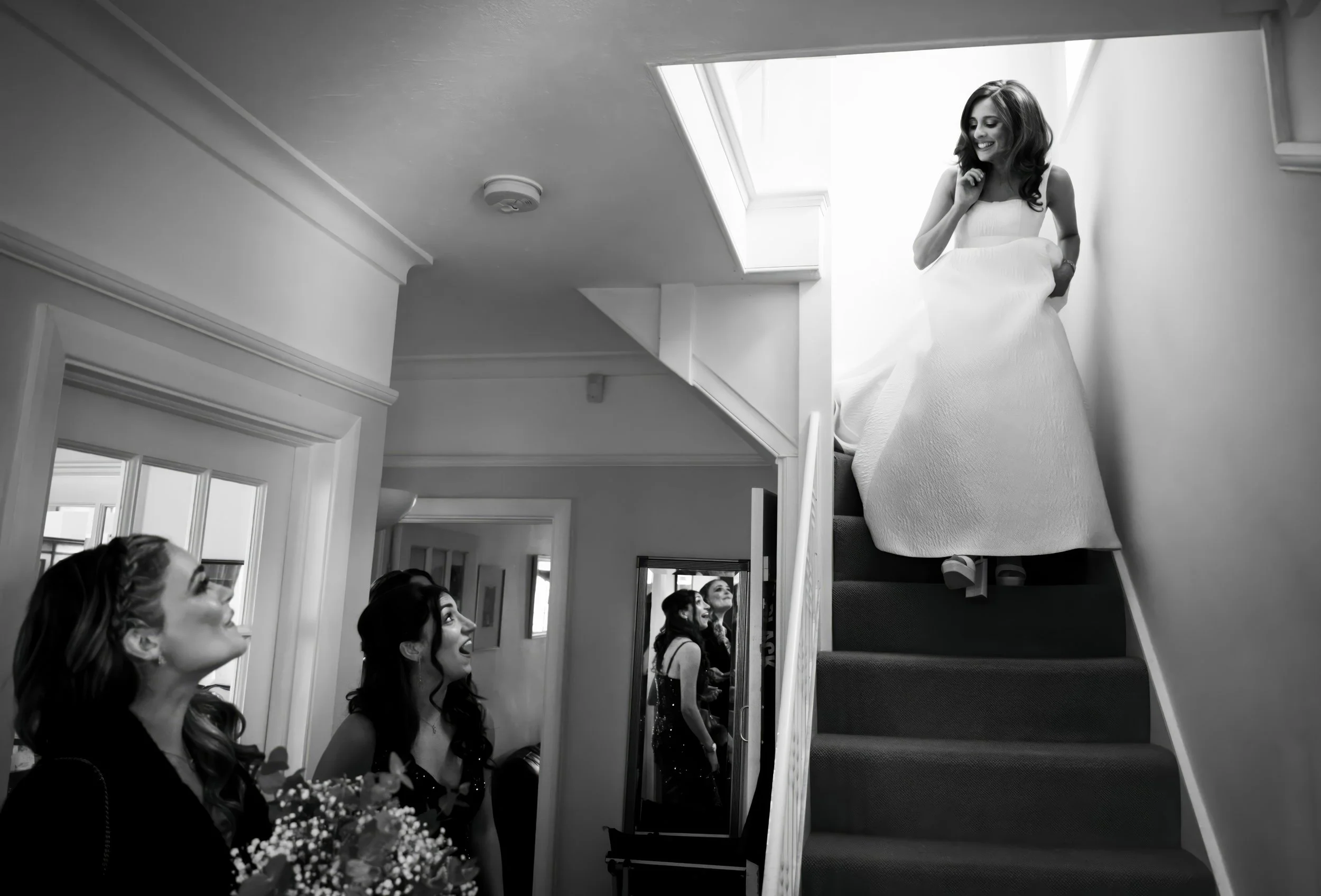 A woman in a wedding dress standing on the staircase, smiling, with three women below looking up at her in a room.