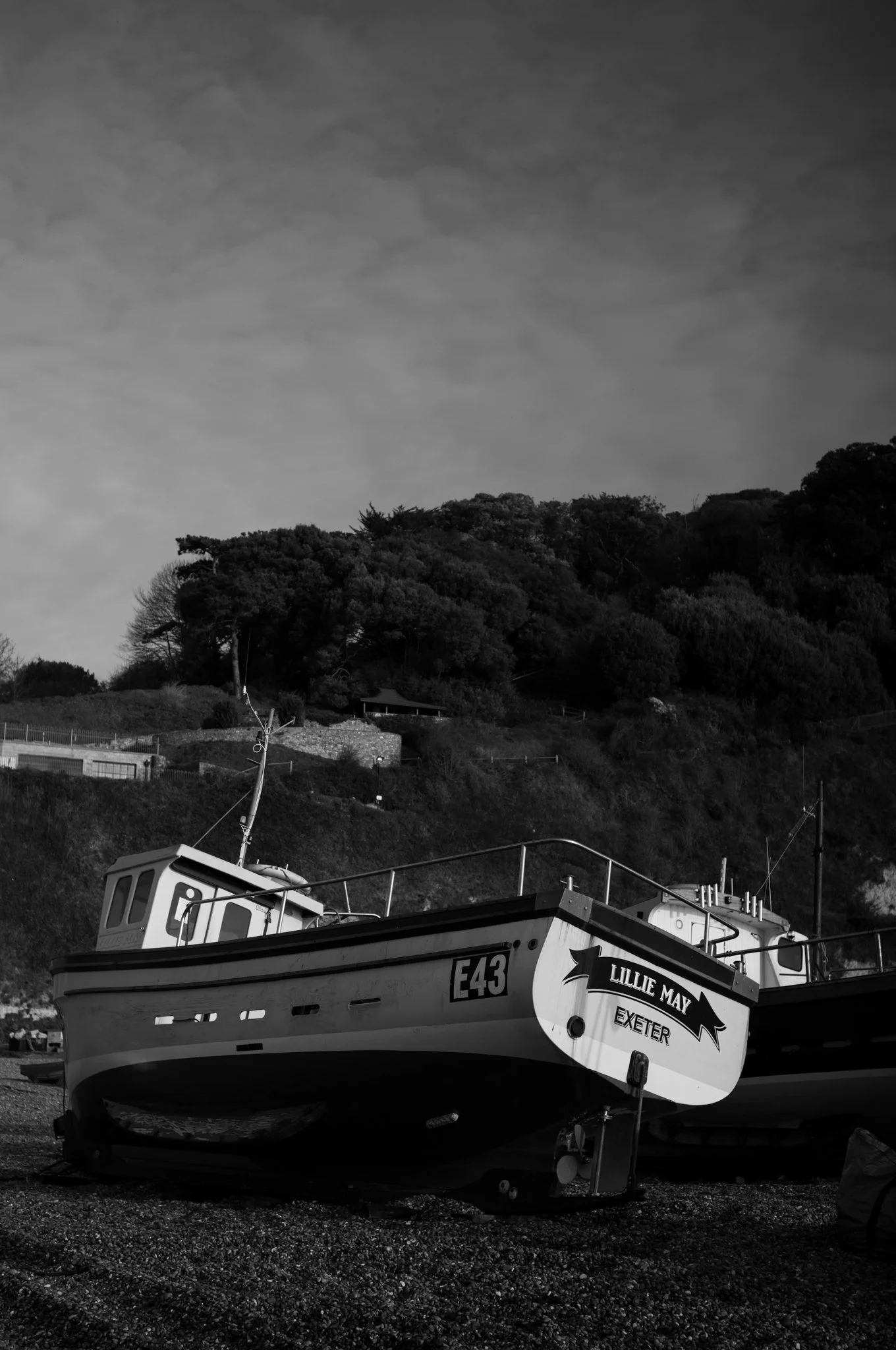 Black and white photo of a boat named "Lillie May" on a dry shore with a hilly, wooded landscape in the background.