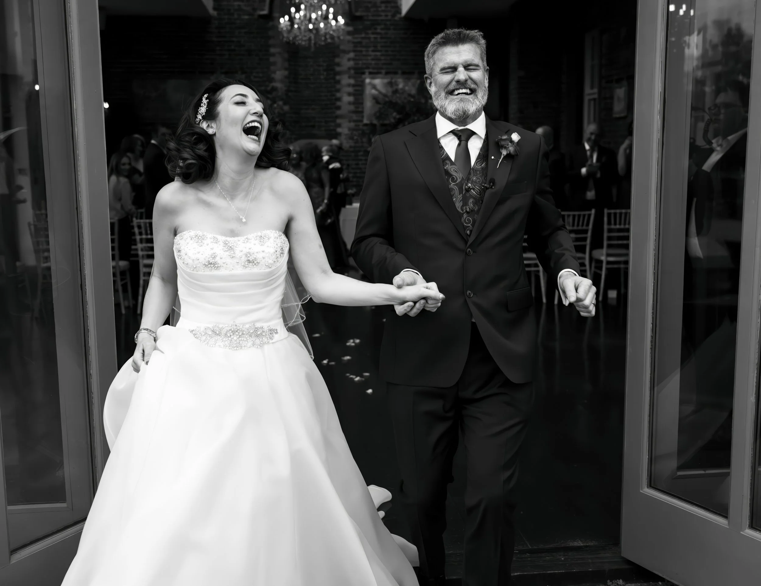 A bride and an older man, possibly her father, are laughing and holding hands as they walk out of a building onto a venue. The bride wears a strapless wedding gown, and the man is dressed in a suit. The background features a chandelier and guests.