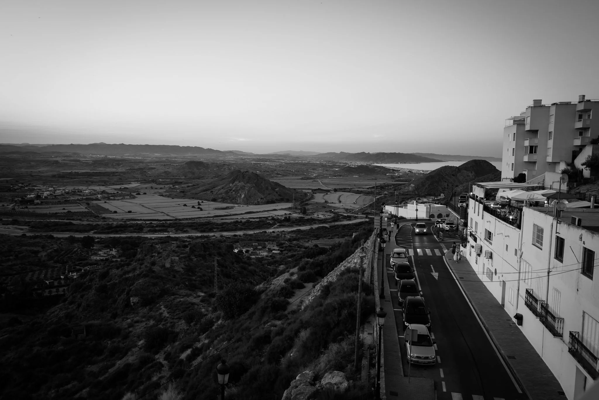 A black-and-white photo of a hillside street with parked cars and apartment buildings on the right, overlooking a valley with mountains in the distance.