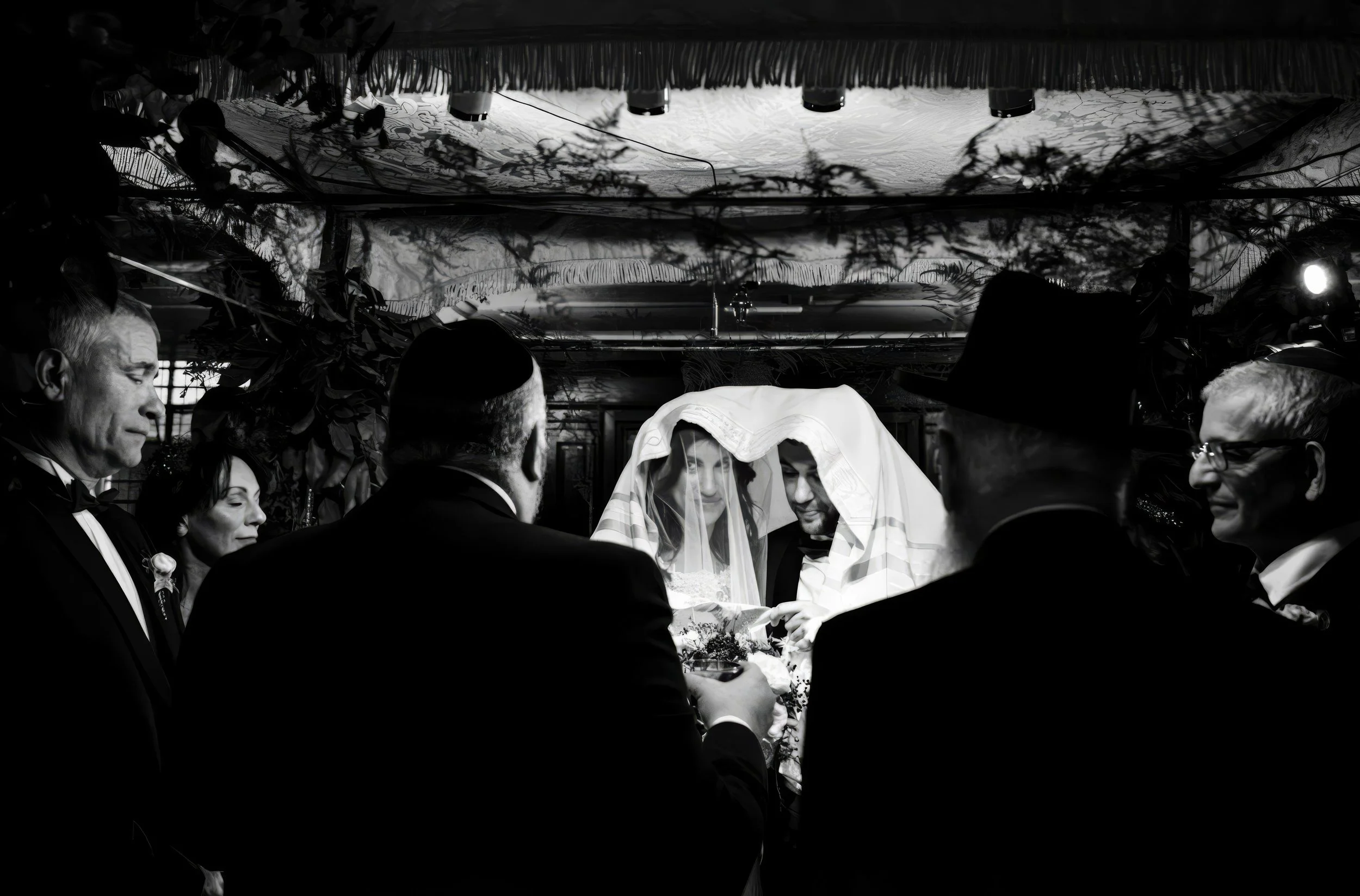 A black and white photograph of a wedding ceremony with the bride and groom under a cloth or veil, surrounded by guests in tuxedos and formal attire.