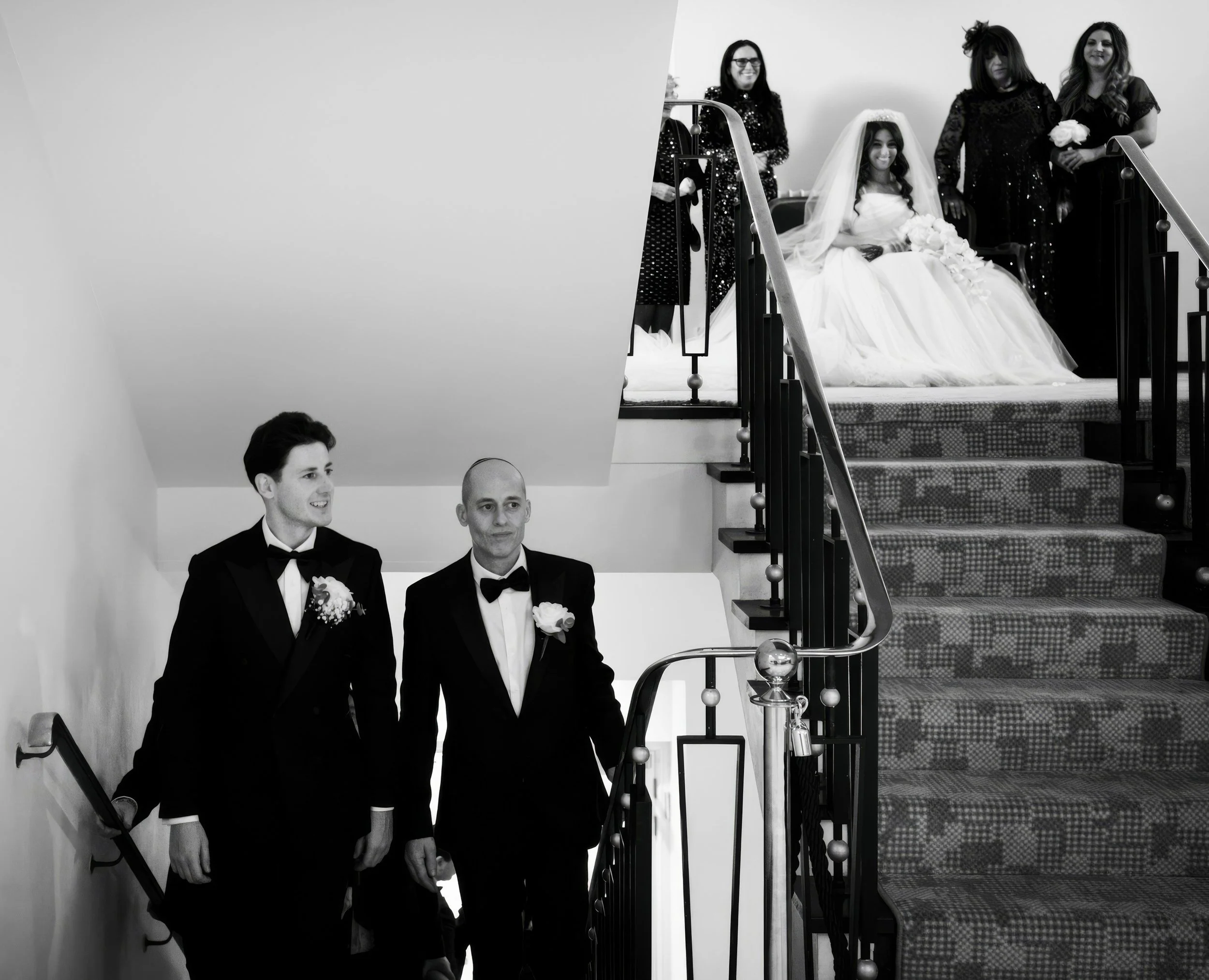 Black and white photo of a wedding scene. Two men in tuxedos walk down stairs, one with a boutonnière, and three women behind a bride seated on the top of a staircase, wearing a wedding dress and veil, holding a bouquet, with two women standing besid