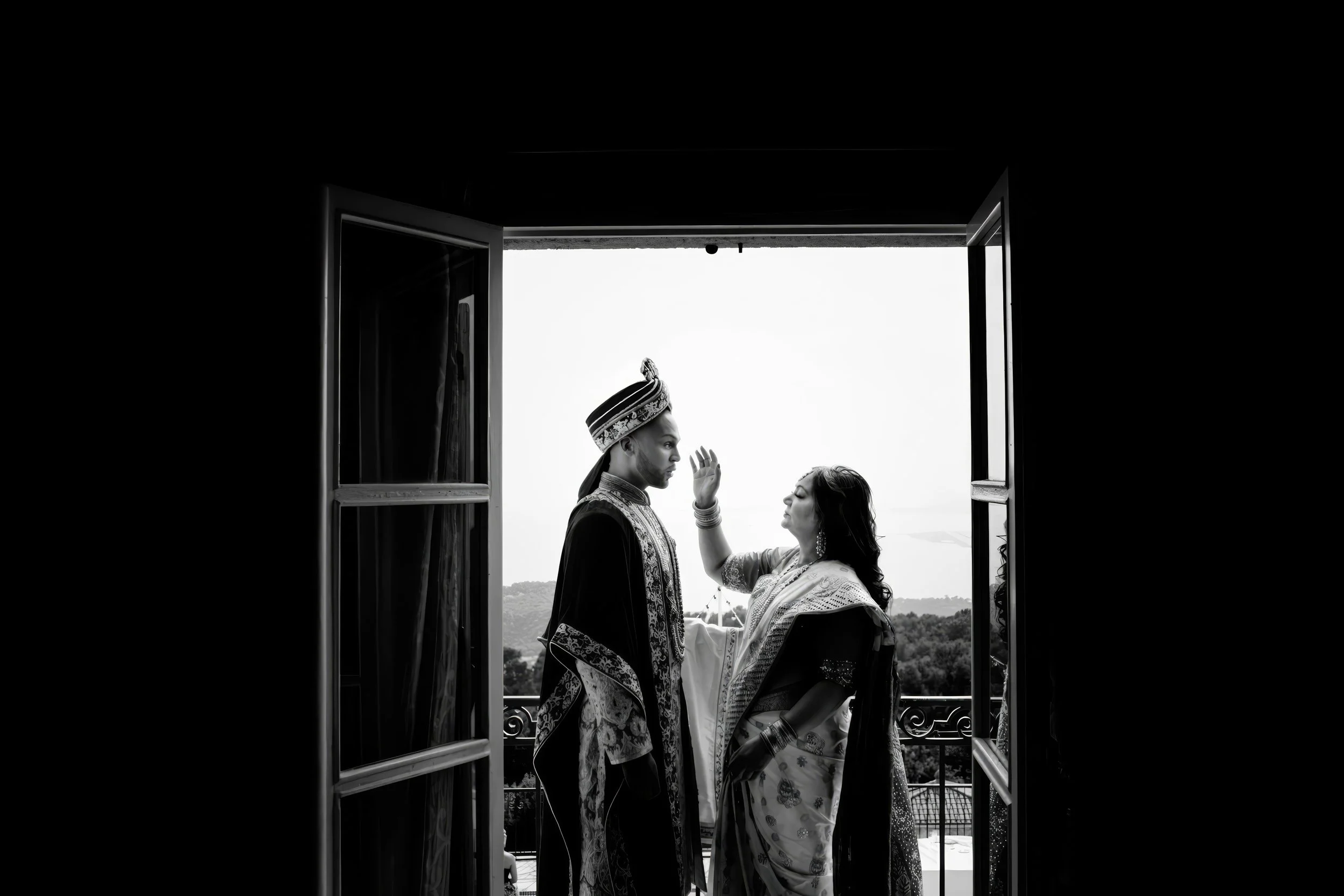 A black and white photo of a couple in traditional Indian wedding attire standing on a balcony, framed by an open window. The woman is touching the man's face, and they are looking at each other with a scenic landscape in the background.