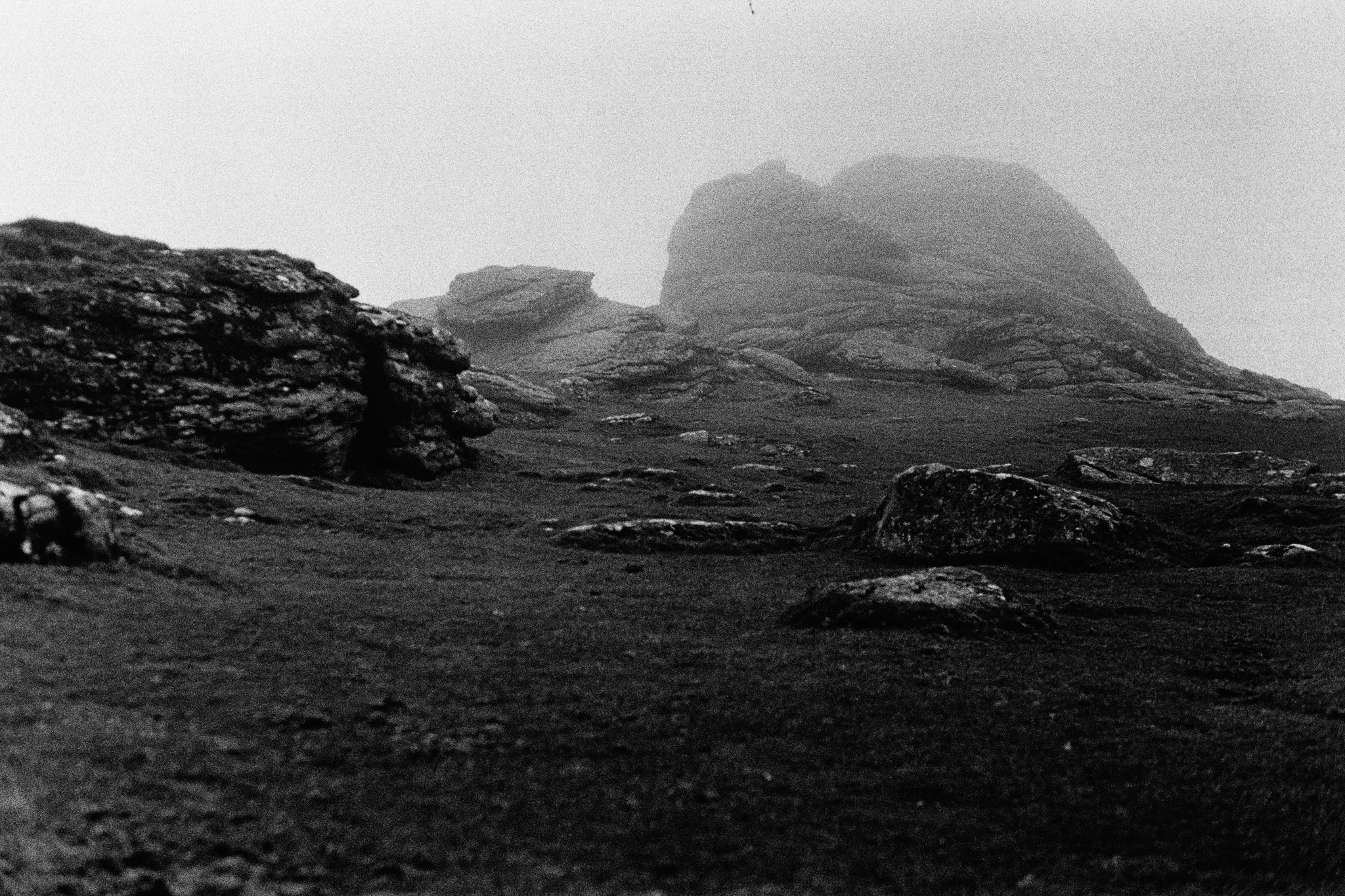 Black and white photograph of rocky terrain with large rock formations in the background, shrouded in fog or mist.