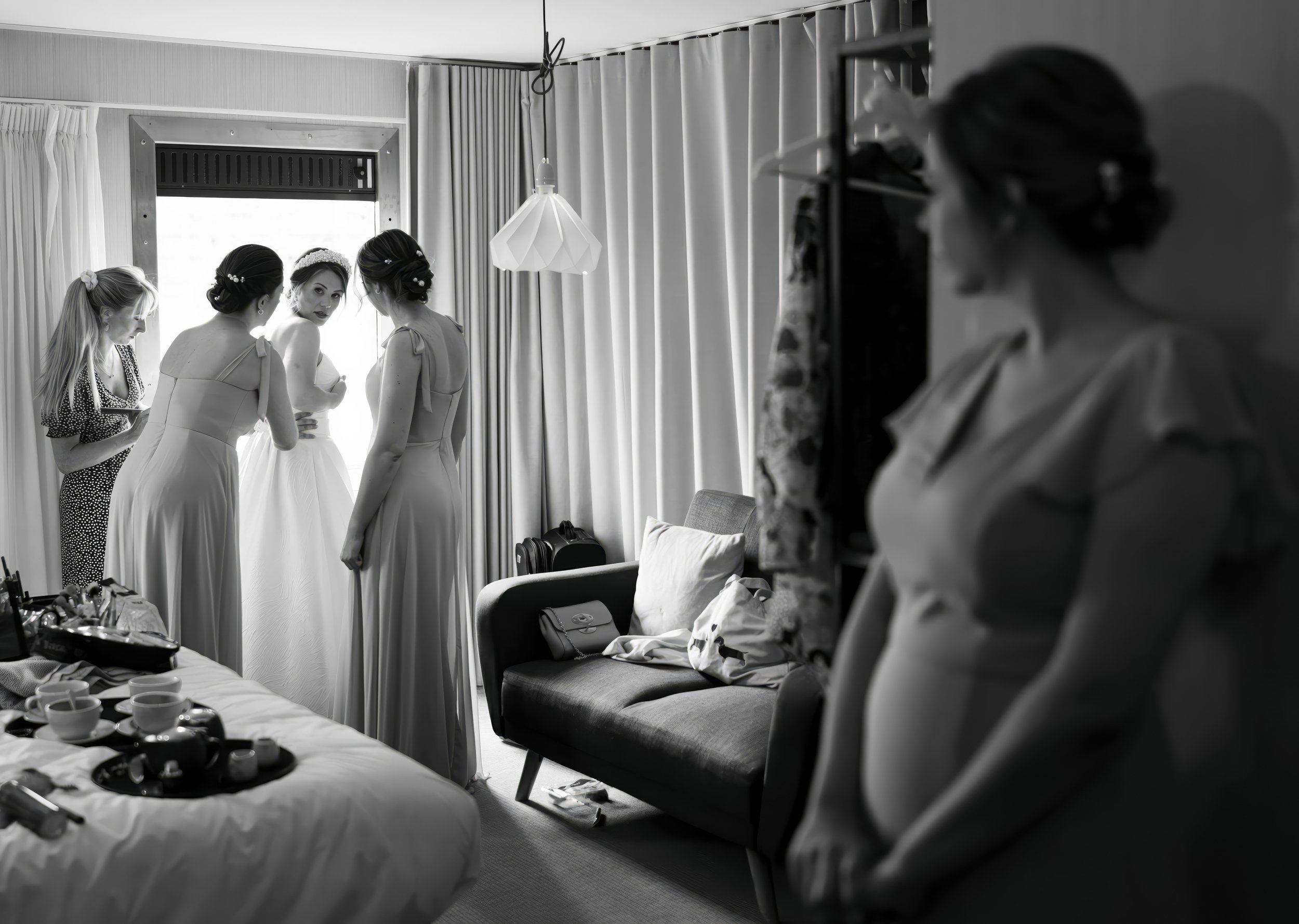 Black and white photo of five women in a bedroom preparing for a wedding. Four women are near a window, engaged in conversation, with one of them wearing a wedding dress. The fifth woman is in the foreground, looking towards the group, standing besid