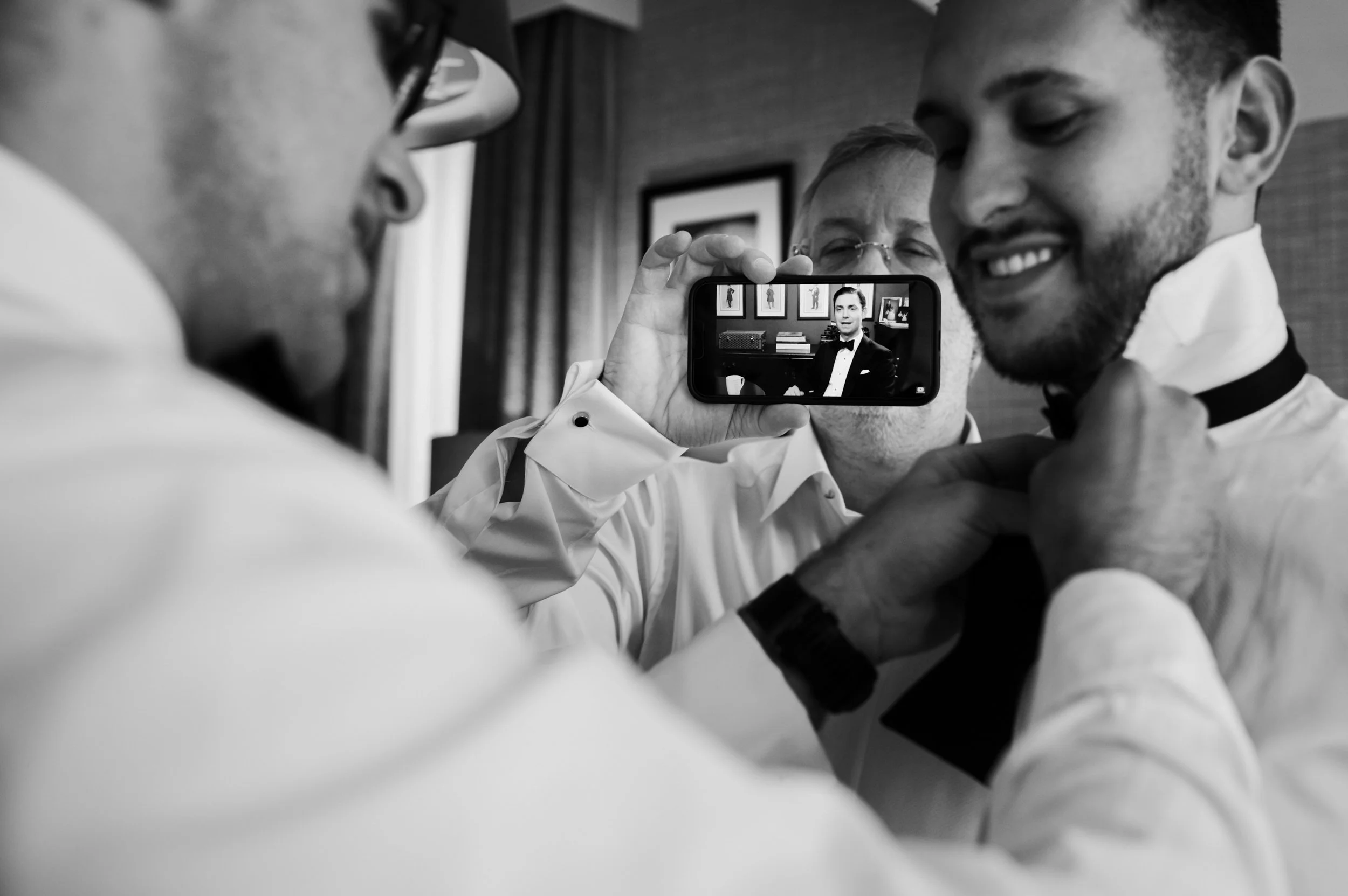 Three men in tuxedos preparing for a wedding, with two adjusting ties and one taking a photo with a smartphone.