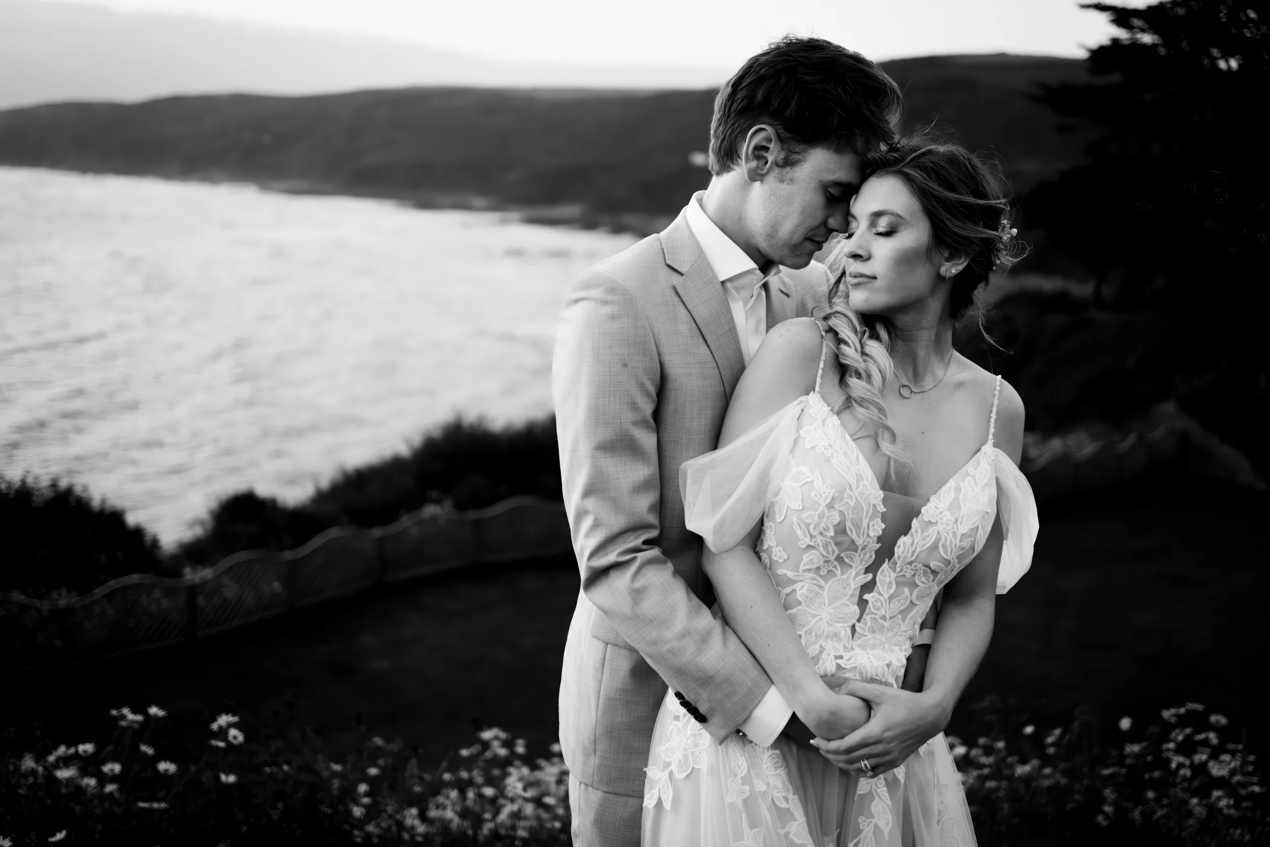 Black and white photo of a couple embracing outdoors near a body of water during sunset, with mountains in the background.