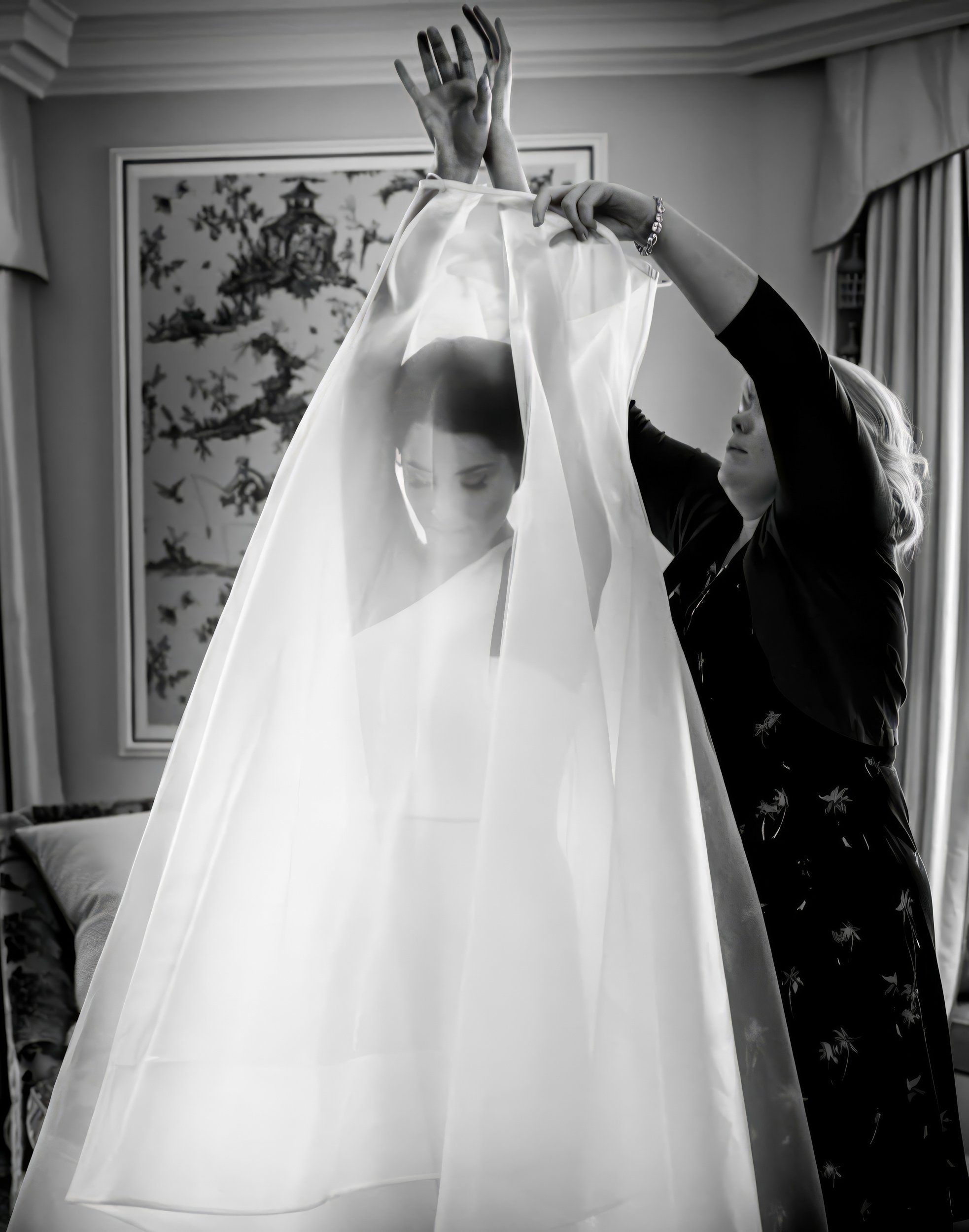 A bride is getting ready, with a woman assisting her by adjusting her veil in a room with curtains and artwork on the wall.