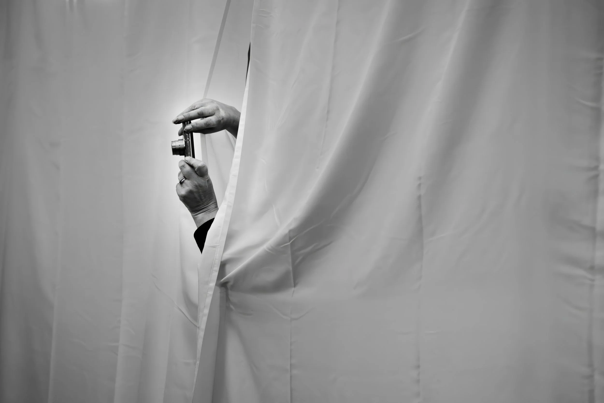 Person holding a camera peeking out from behind a curtain in black and white.