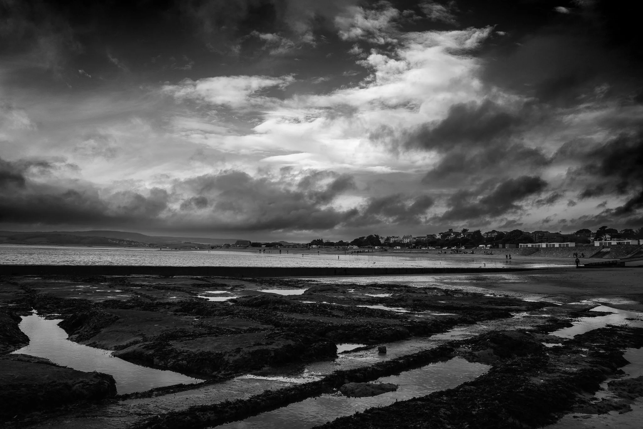 A black and white photo of a beach with rocky tide pools in the foreground, calm water in the middle ground, and a distant shoreline with houses and trees under a cloudy sky.