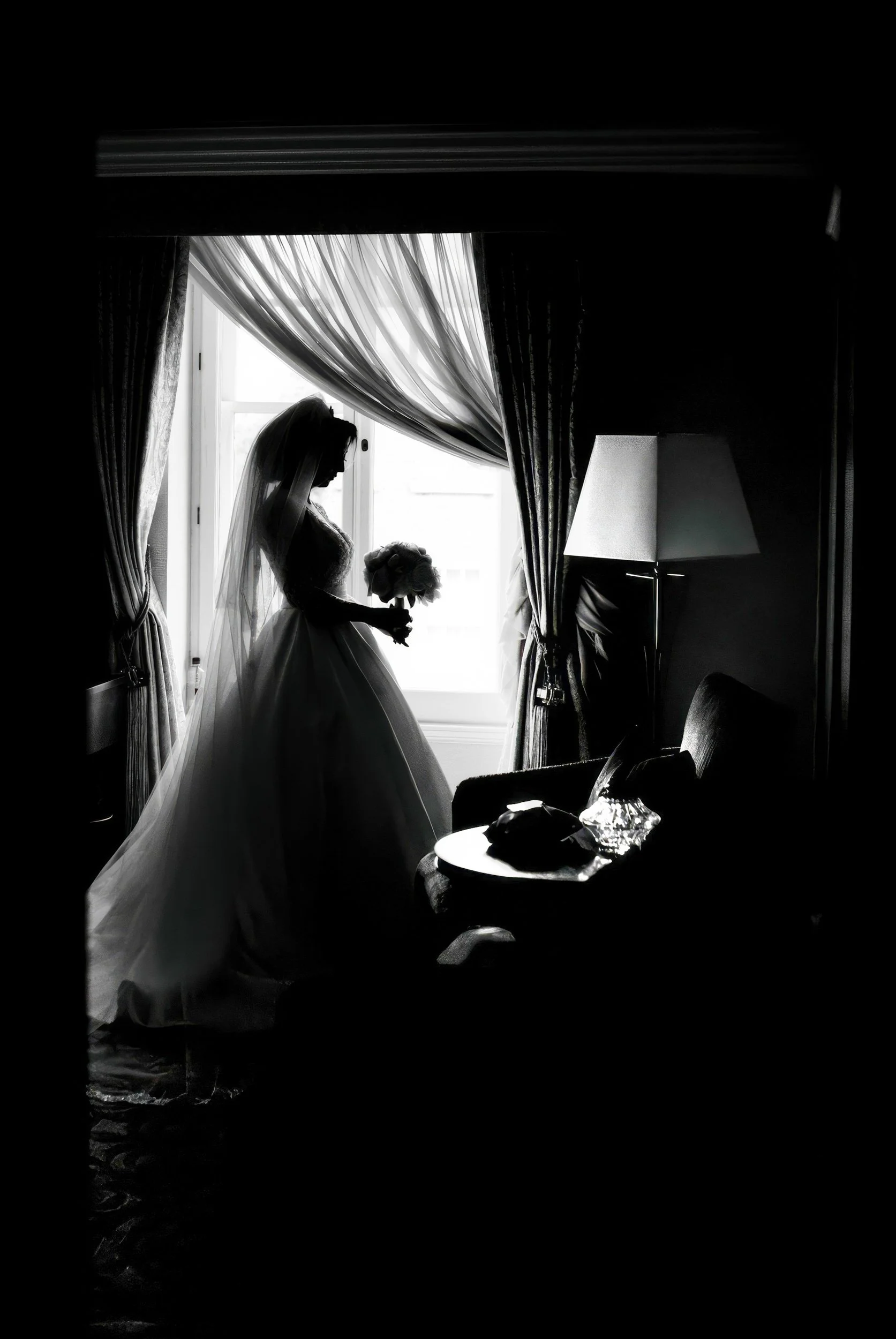 Silhouette of a bride holding a bouquet of flowers, standing by a window with curtains in a dimly lit room.