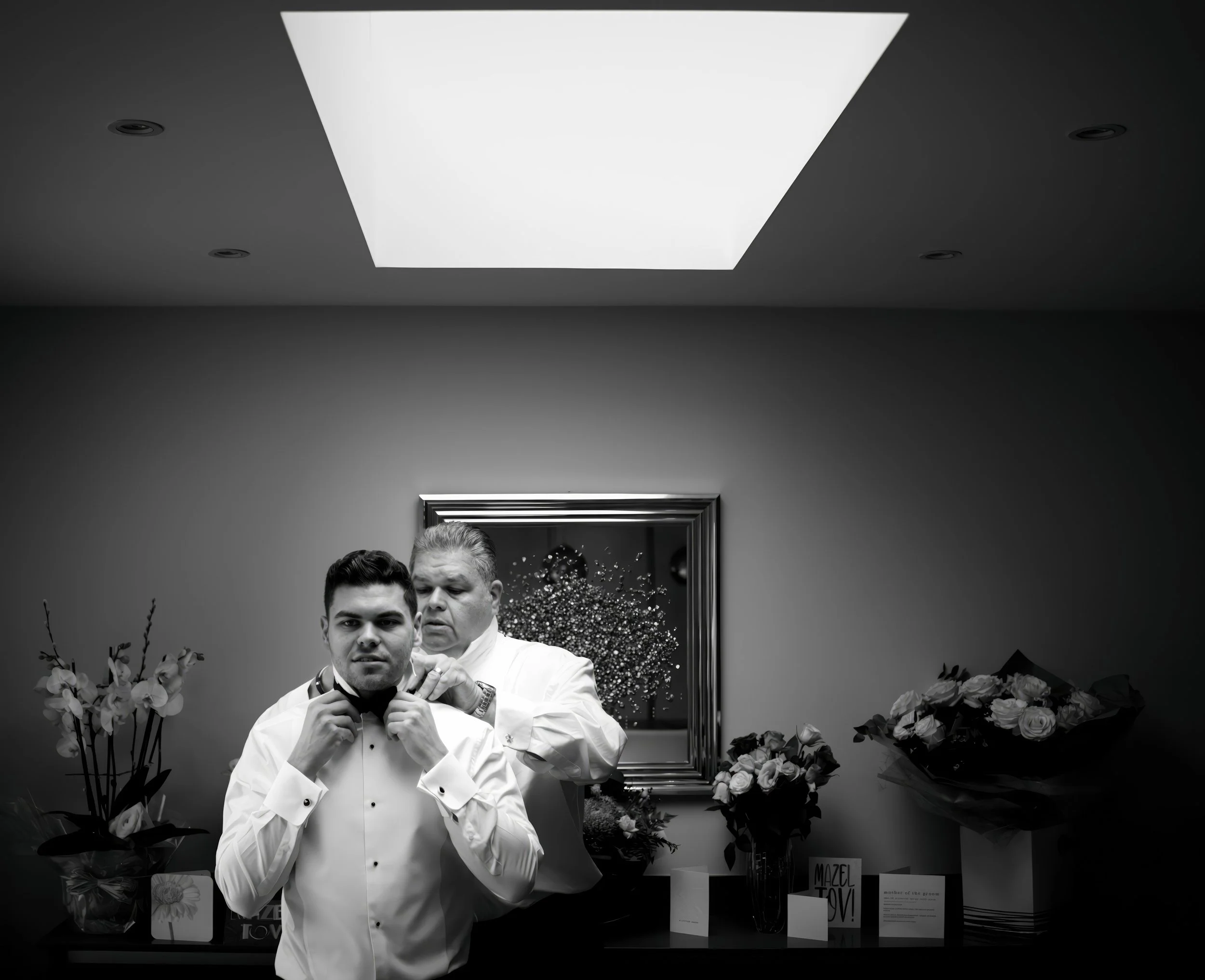 Two men in formal attire, one adjusting a bow tie and the other helping, standing in a room with flower arrangements and a mirror behind them.