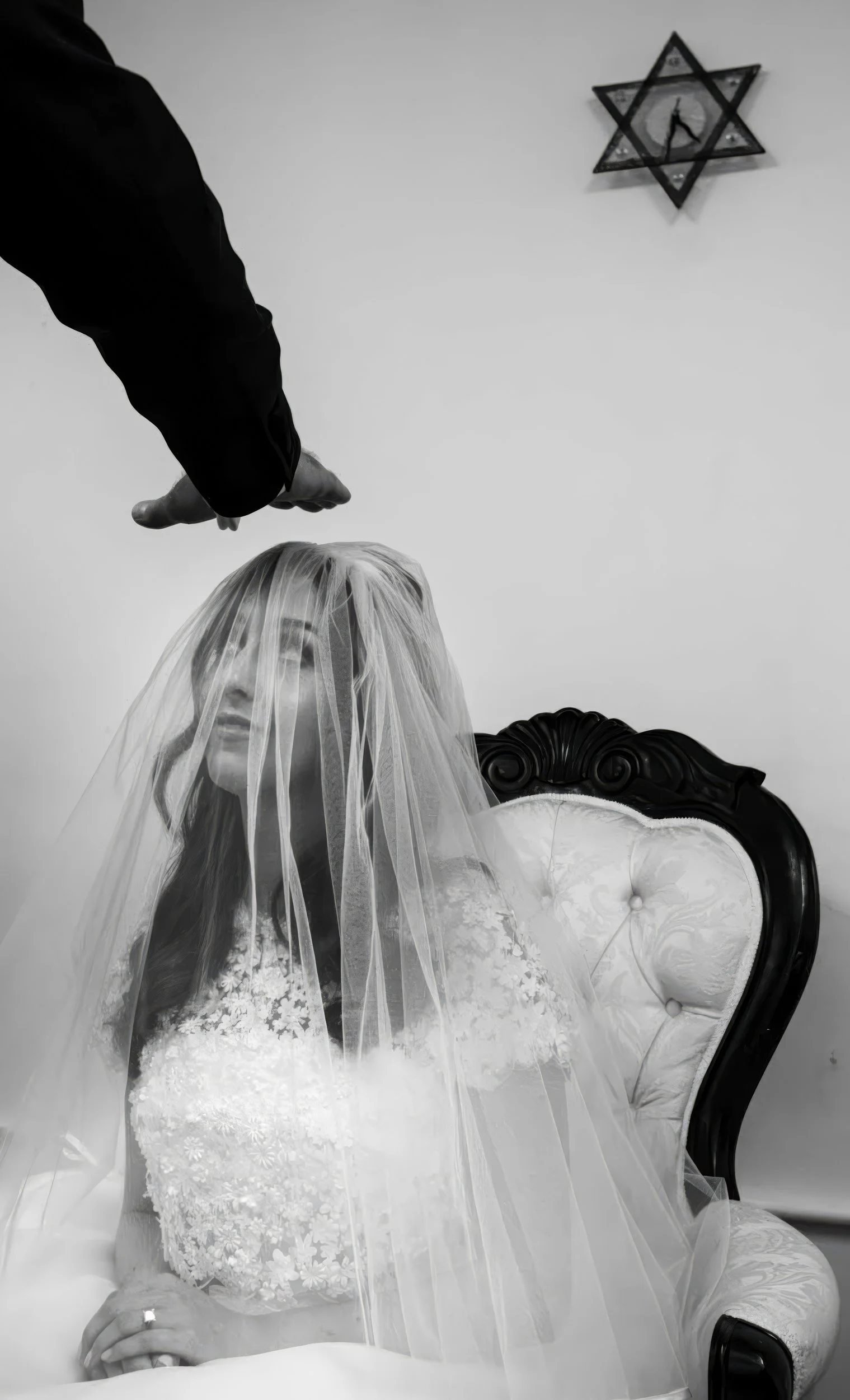 A bride sitting on a vintage chair with a veil covering her face, and a hand pointing at her from above. A Star of David decoration hangs on the wall in the background.