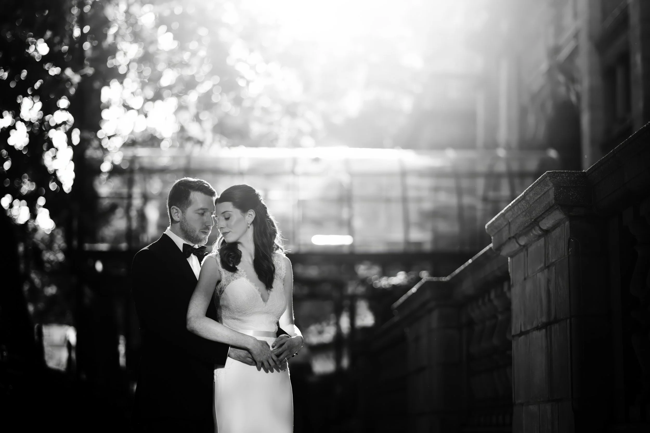 Black and white photo of a bride and groom embracing outdoors in front of a stone railing with sunlight filtering through the trees.