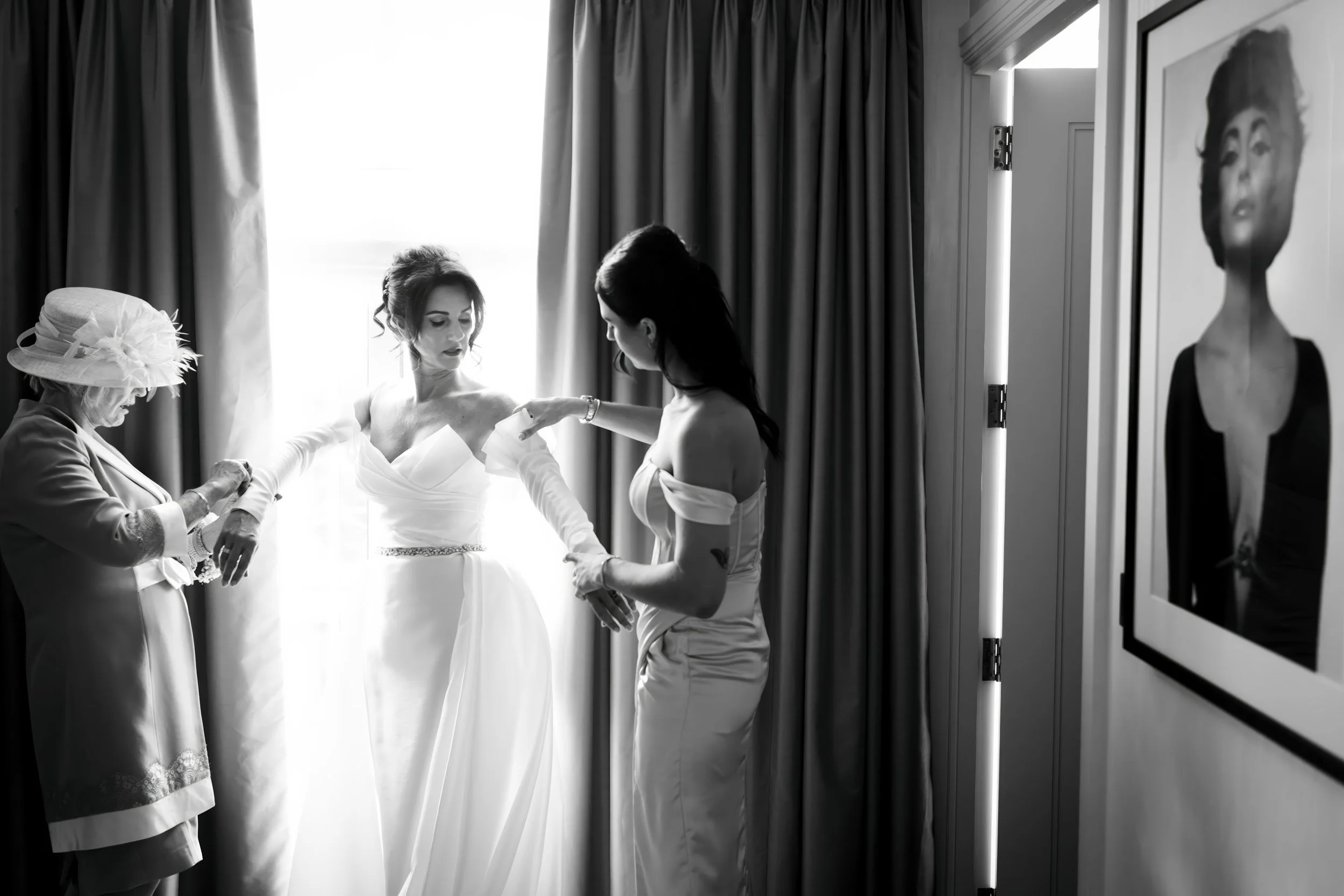 A woman in a wedding dress is being assisted by another woman in a strapless dress, while an older woman in a hat helps with her accessories in a room with curtains and a framed black-and-white portrait on the wall.