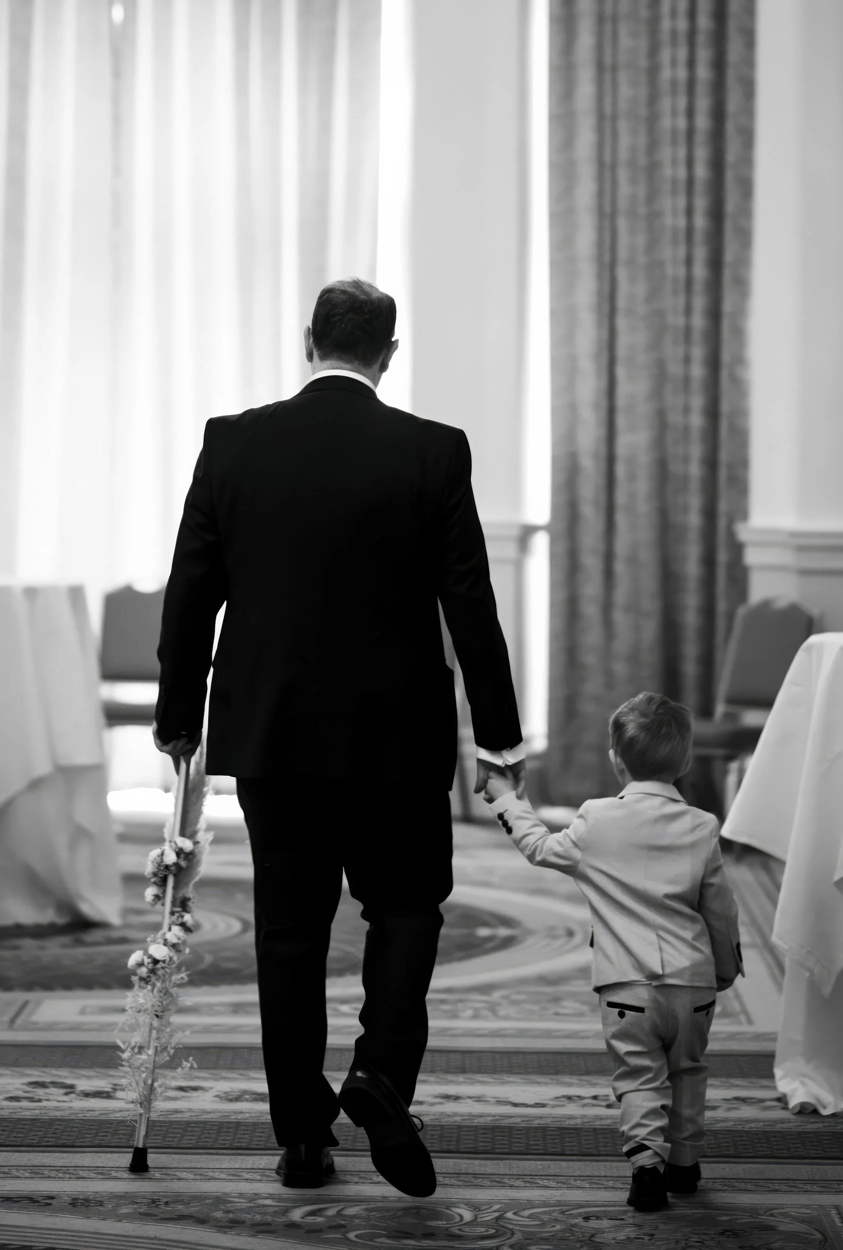 Man in a dark suit holding a child's hand while walking in a room with carpeted floor and large curtains