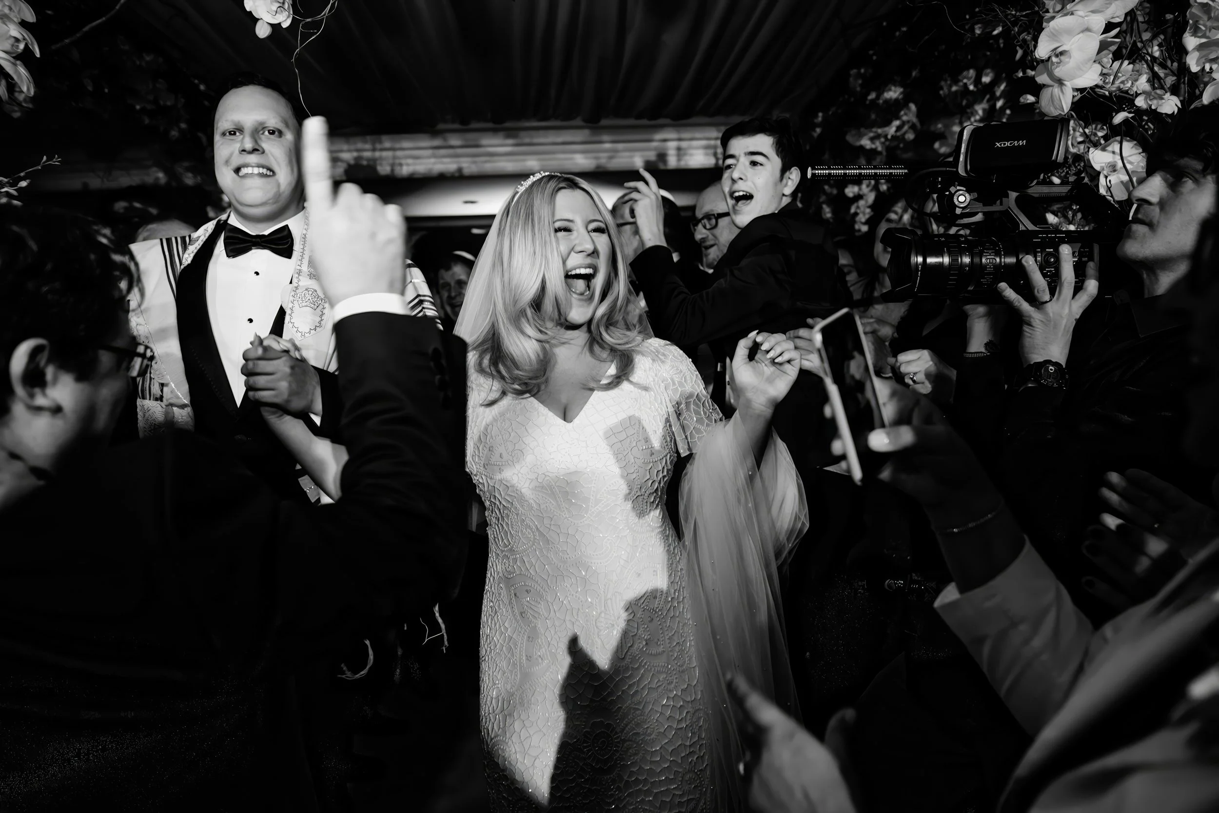 Black and white photo of a joyful woman in a wedding dress surrounded by people celebrating, including a man in a tuxedo and others taking photos and videos with cameras and smartphones.