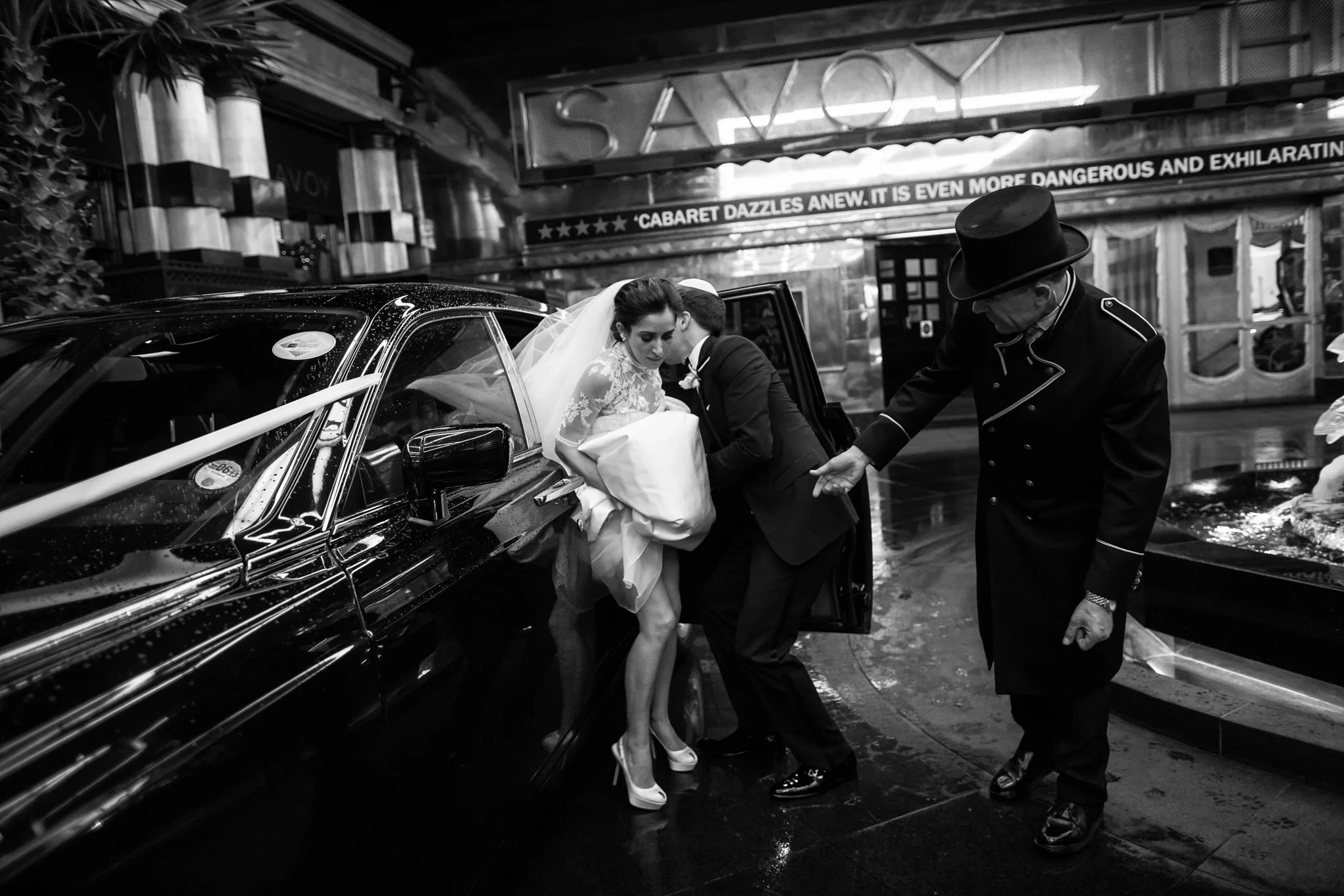 A bride and groom step out of a black limousine, assisted by an attendant wearing a top hat, on a rainy city street at night. The bride is wearing a lace wedding dress and high heels, and the groom is in a tuxedo. The background shows a casino and br