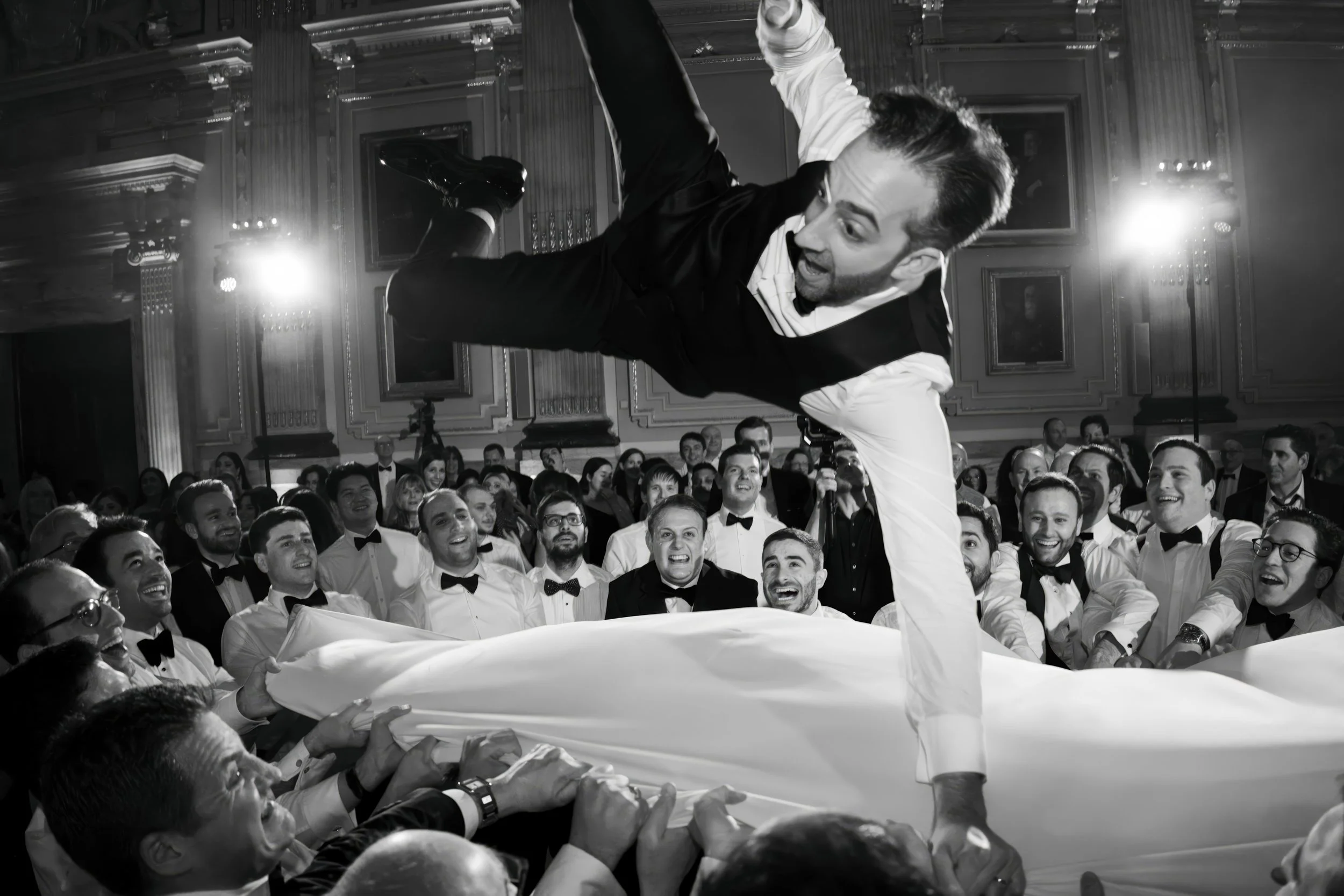 Man performing a show or dance move on a sheet held by a crowd at a formal event in a grand hall, with many people dressed in tuxedos watching and smiling.
