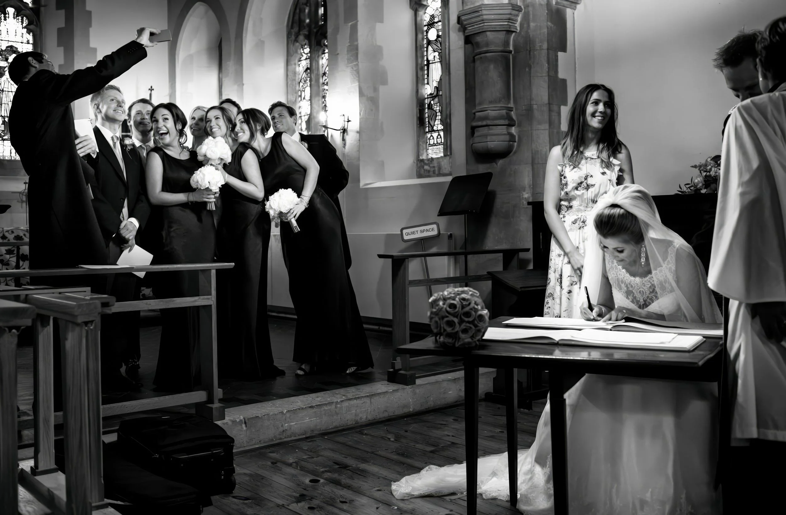 A bride signing a book during her wedding ceremony in a church, with a group of bridesmaids and groomsmen taking a group selfie in the background.