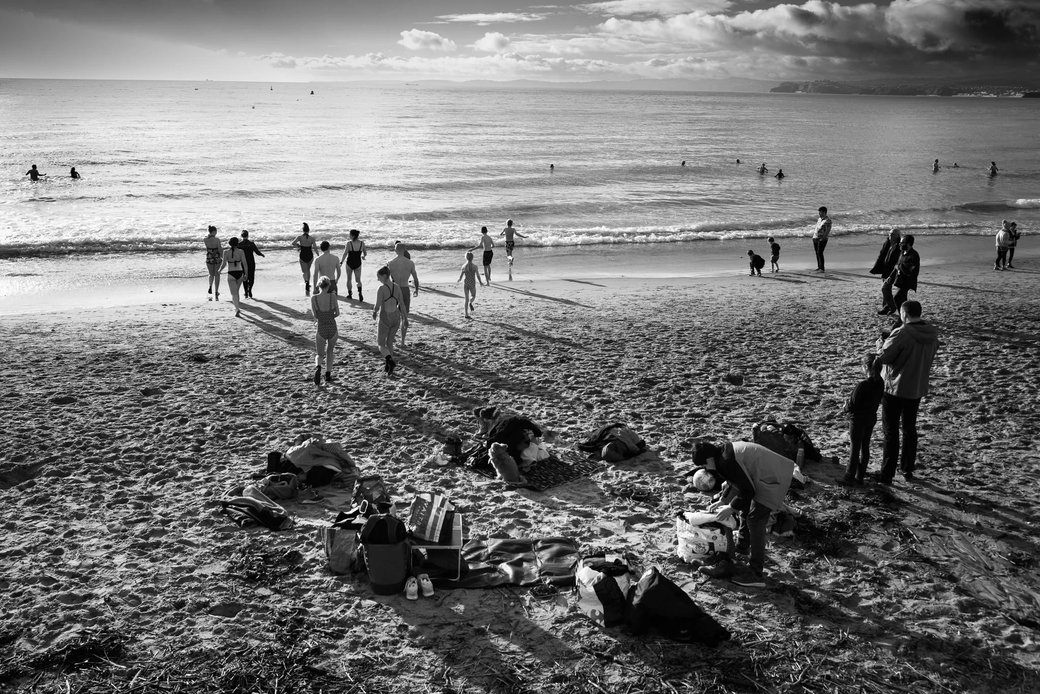 A black and white photo of a beach scene with people swimming and walking along the shore. Surfboards are visible in the water, and groups of people are sitting or standing on the sand, some with towels and bags.